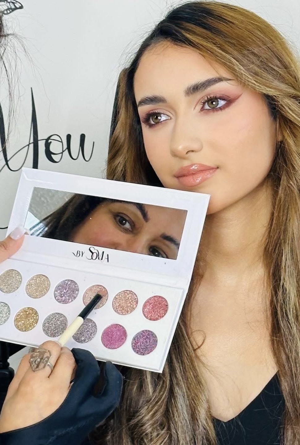 Woman with makeup looking at a mirror palette, makeup artist applying eyeshadow, close-up of face and eye makeup, neutral background.
