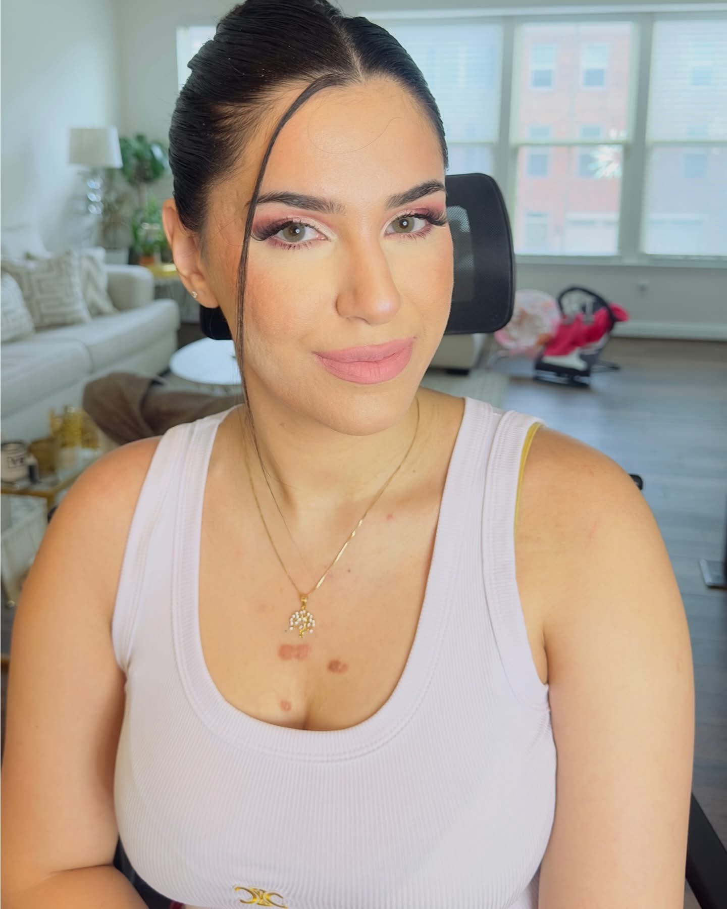 Young woman with dark hair in a bun, wearing a white tank top, smiling in a bright room with large windows.