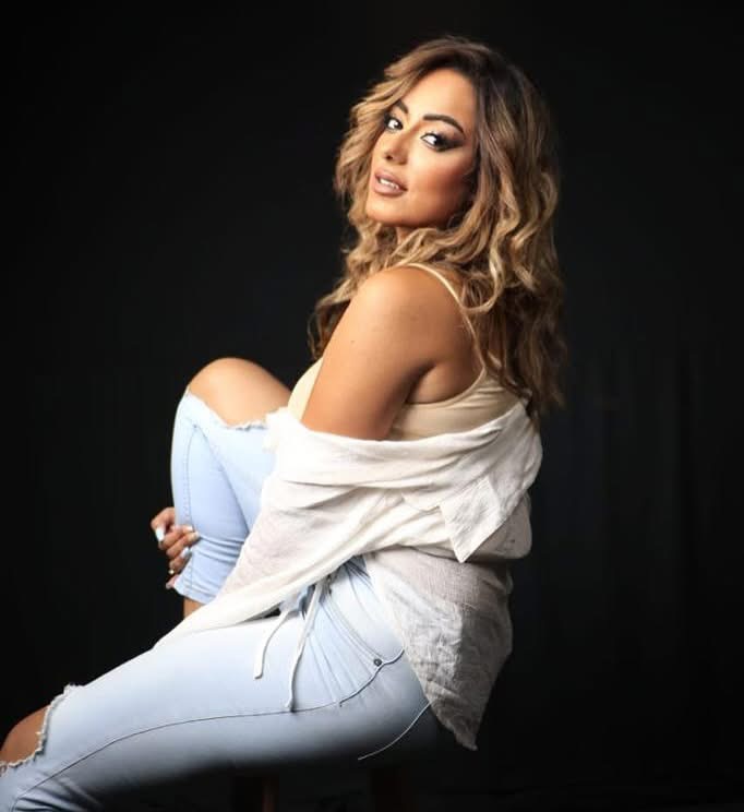 Woman with wavy hair sitting against a black background, wearing a white off-shoulder top and ripped jeans.