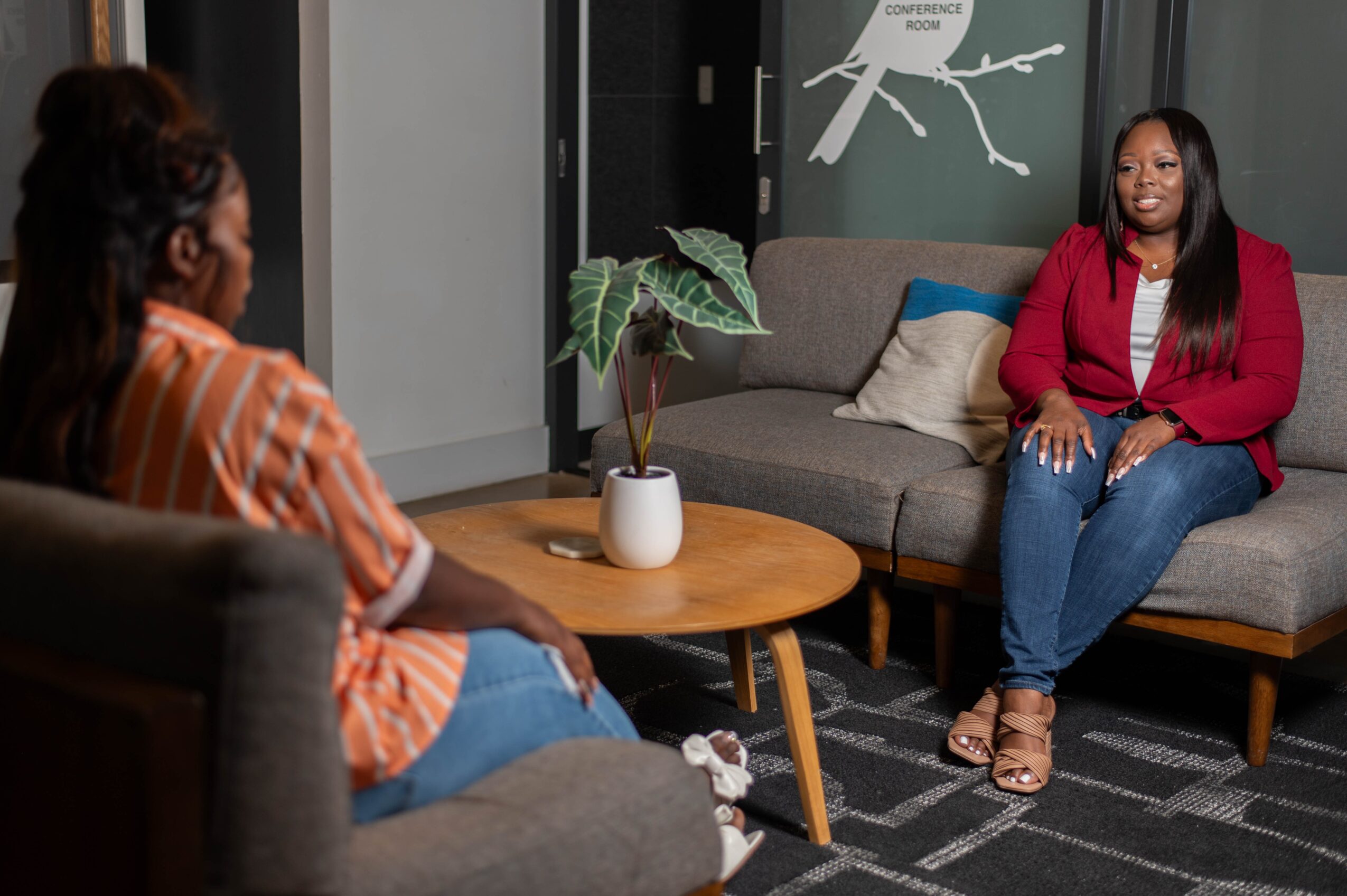 Two women sit in a living room, talking, with a sofa, a round wooden table, and a potted plant between them.