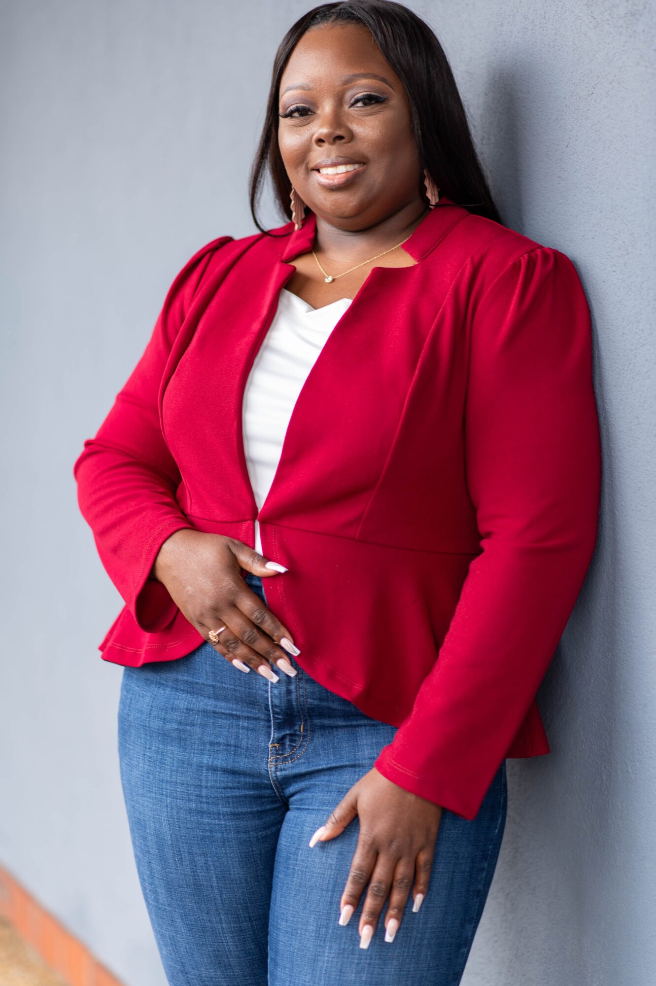 Woman in red blazer and jeans leaning against a wall, smiling, with a light gray background.