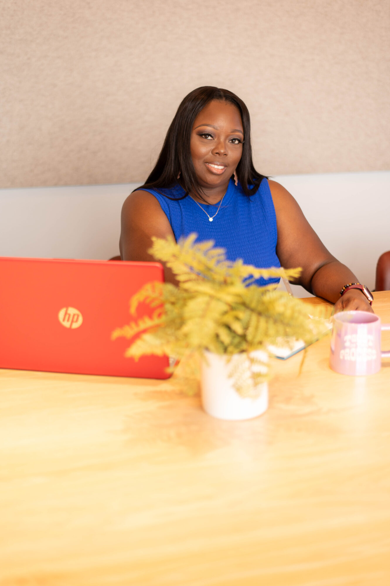 Woman in blue top sitting at table with laptop, plant, and candle, smiling, beige wall background.