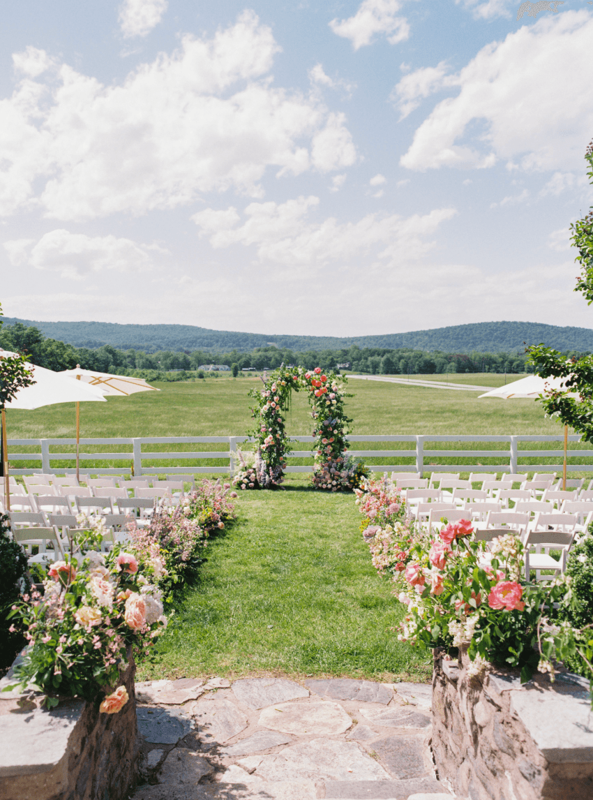 Outdoor wedding setup with floral arrangements, chairs, and an arch in a green field under a partly cloudy sky.
