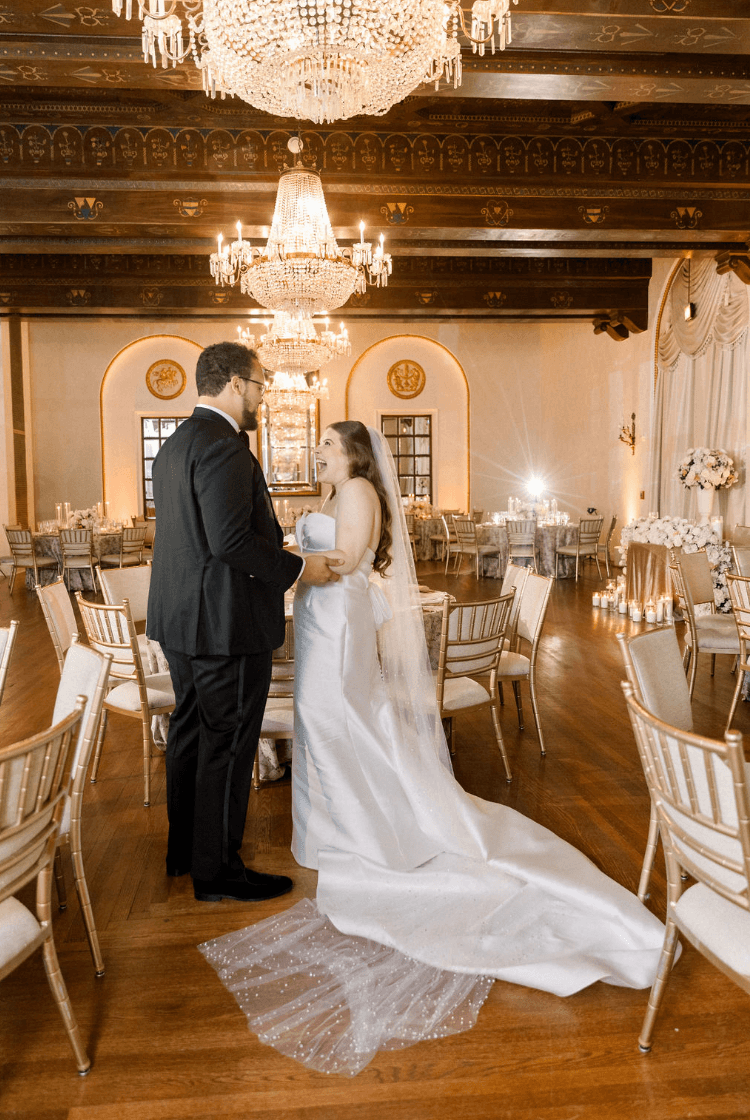 A bride and groom stand facing each other in a decorated wedding venue with chandeliers and round tables, holding hands.