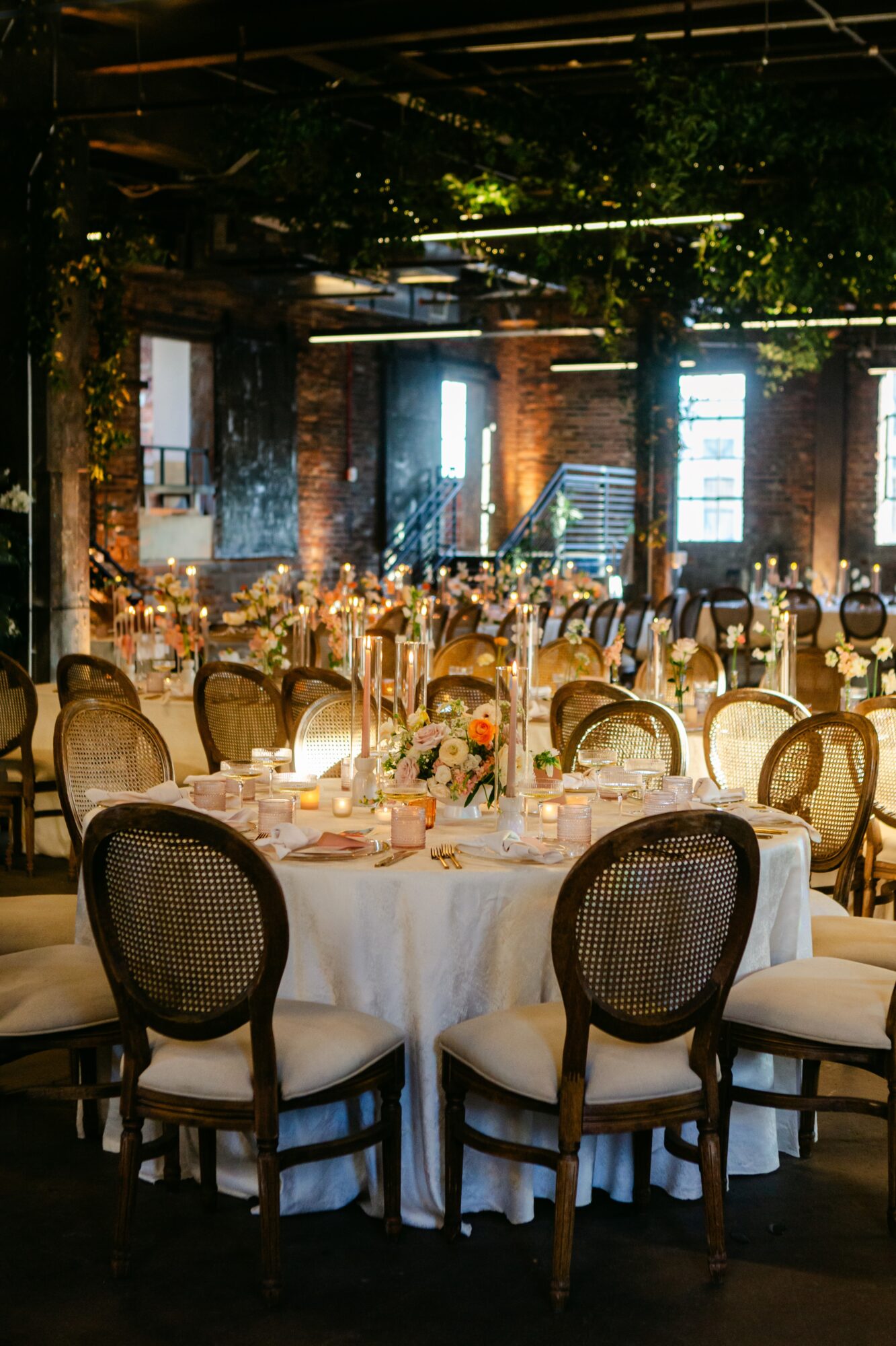 Elegant banquet table set with floral centerpiece, surrounded by chairs, in a decorated indoor venue with brick walls and large windows.