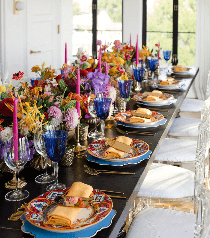 Long dining table set with colorful floral arrangements, blue glassware, and place settings with napkins and plates, in a bright room.