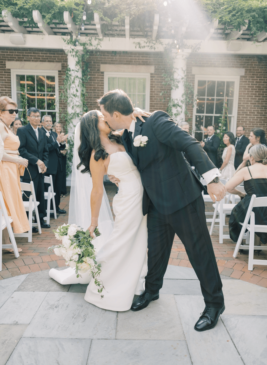 Groom dips bride for a kiss outdoors, surrounded by guests and greenery, during a wedding celebration.