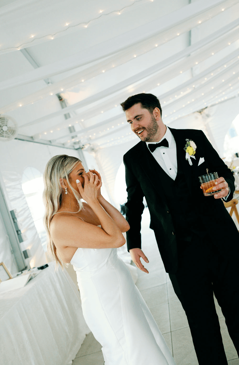 A bride and groom laughing and smiling at each other during their wedding reception, with string lights overhead.