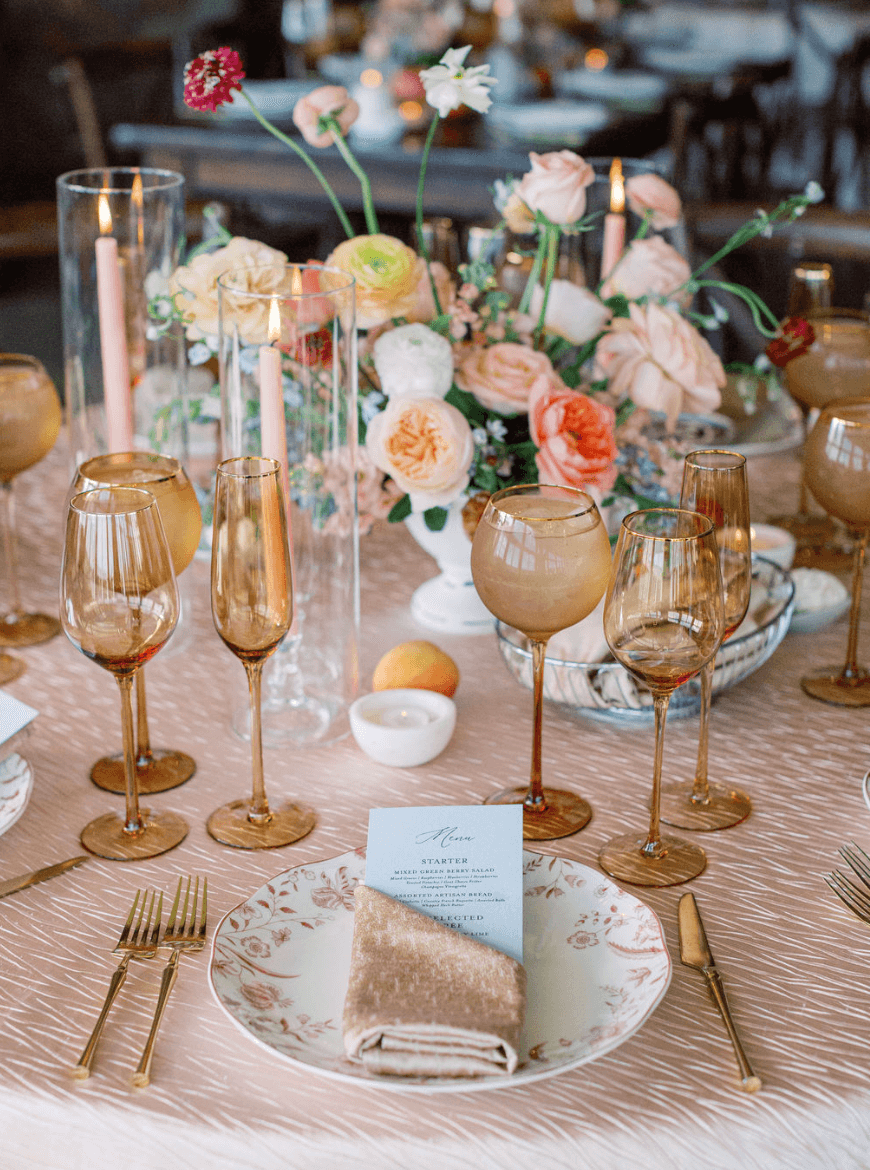 Elegant table setting with a floral centerpiece, gold-rimmed plates, glasses, and a slice of cake on a plate.