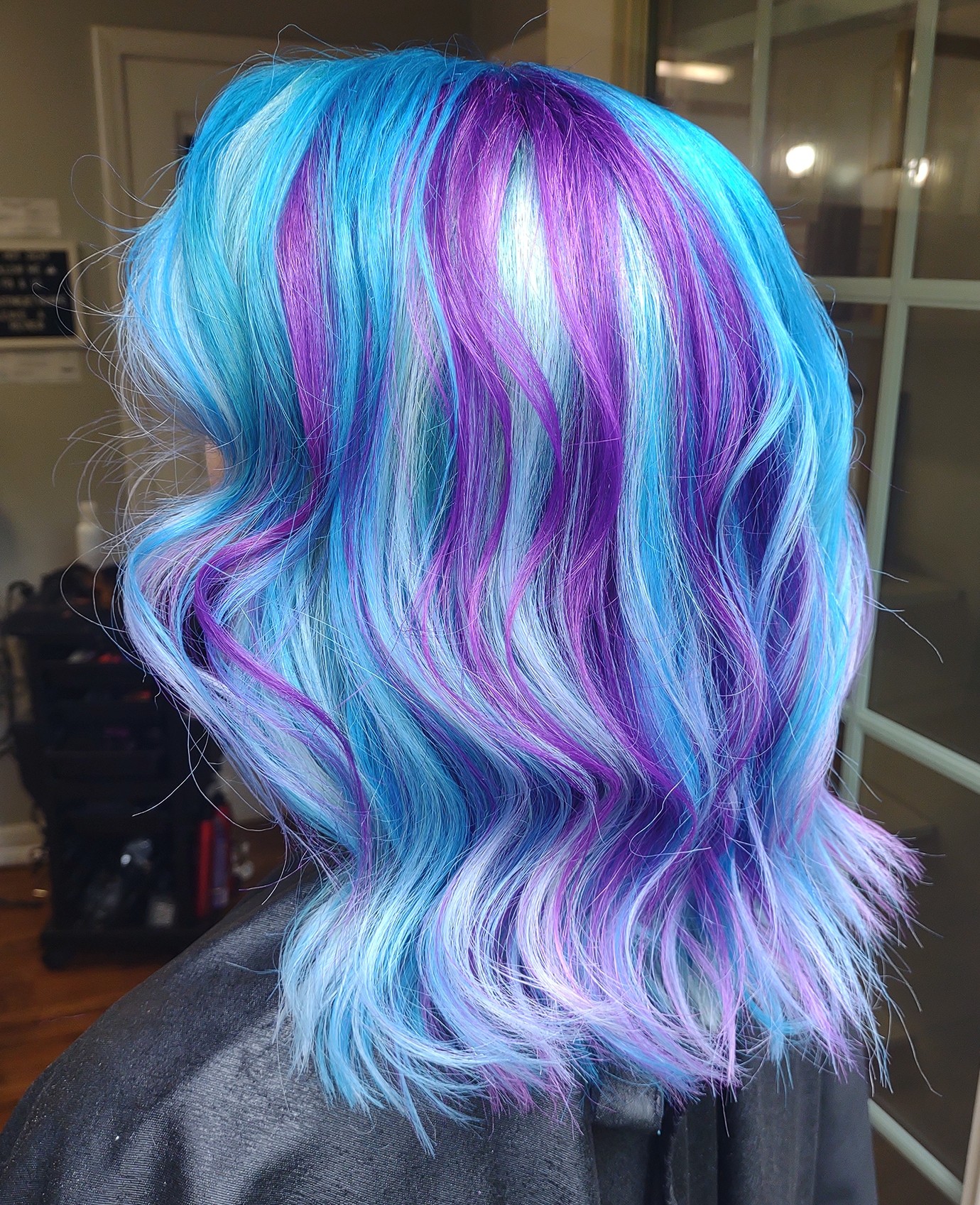 Person with wavy, multicolored hair in shades of blue, purple, and white, styled in loose waves, indoors.