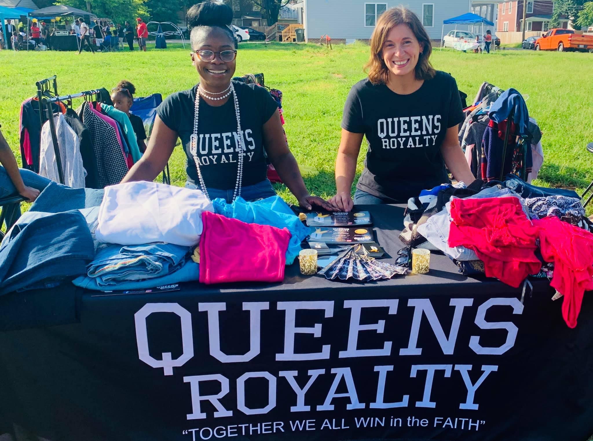 Two smiling women stand behind a table with Queens Royalty banner, selling clothes outdoors on a sunny day.