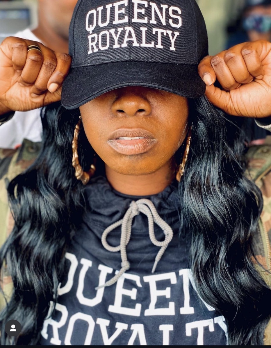 Woman wearing a black cap with white text, holding the cap's brim, with long black hair and hoop earrings.
