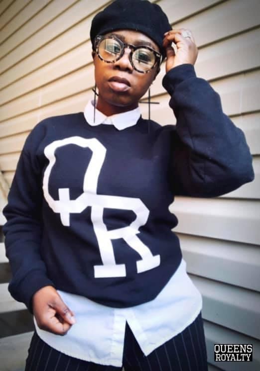 Person wearing glasses, black beanie, black sweater with white design, white shirt underneath, standing against beige horizontal siding.