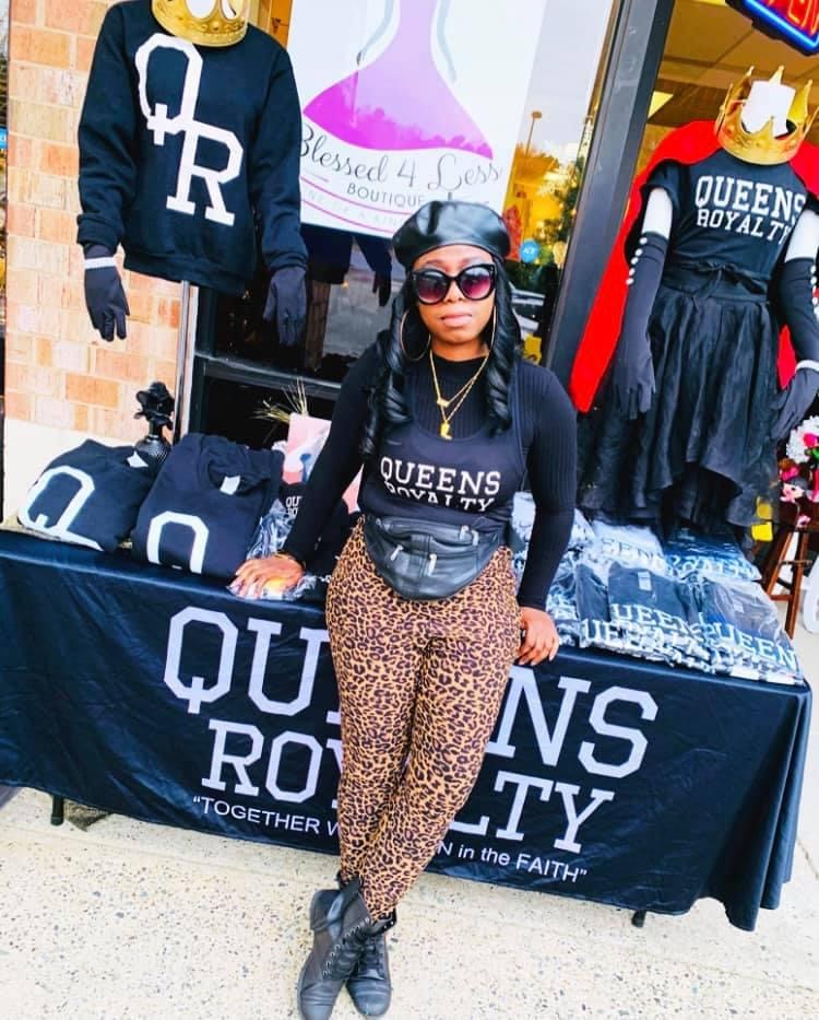 Woman with sunglasses and long braids standing in front of a Queens Royalty booth, wearing leopard print pants and a black top.