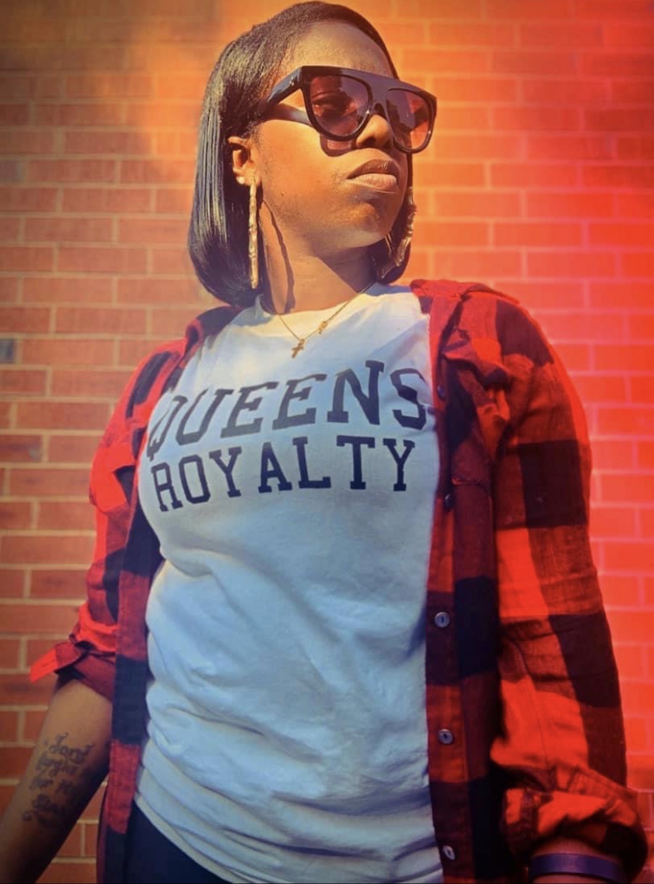 Young woman wearing sunglasses, a white T-shirt with 'QUEENS ROYALTY' text, and a red plaid shirt, standing outdoors against a brick wall.
