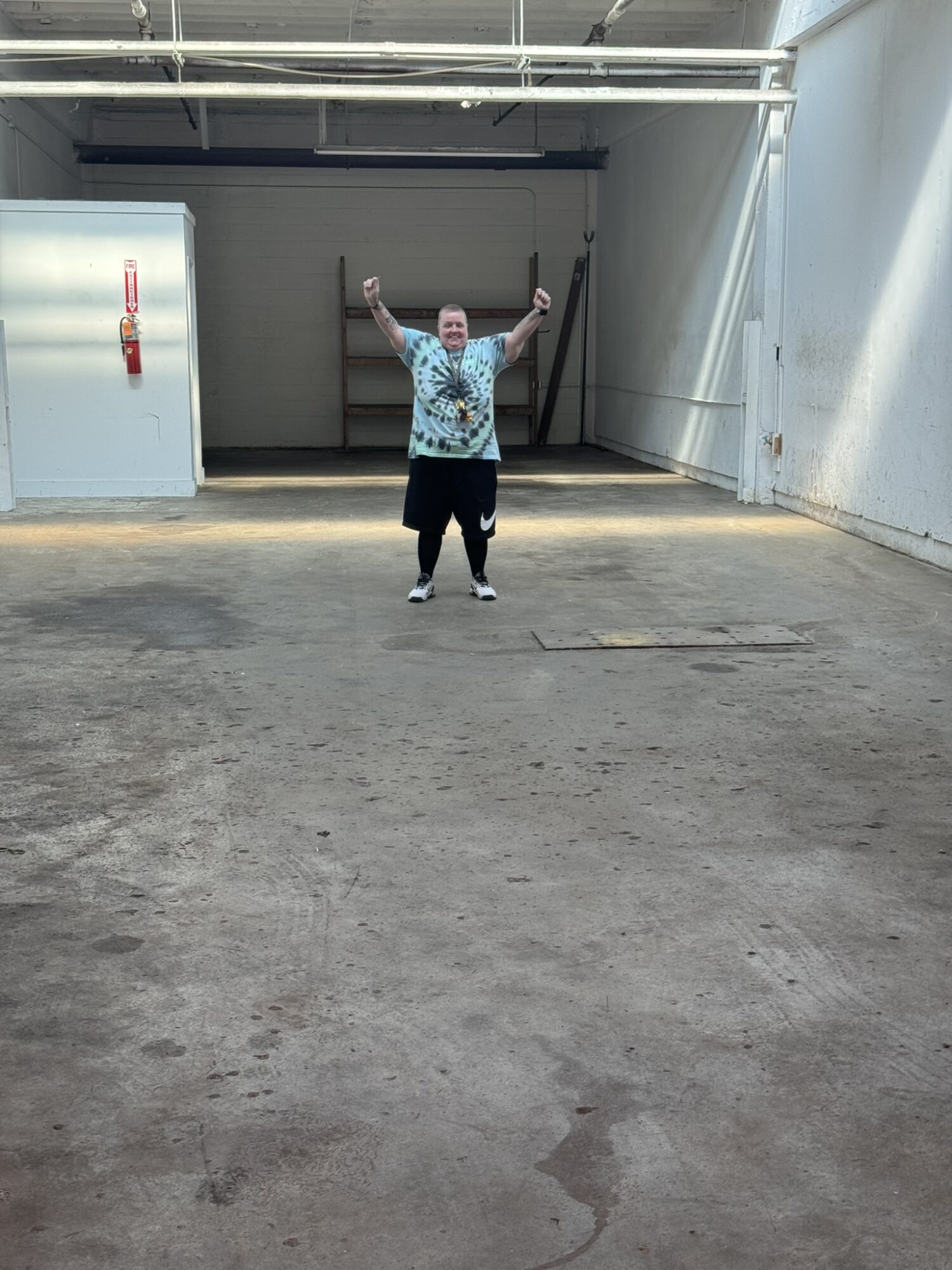 Person standing in an empty parking garage with arms raised, wearing a patterned shirt and dark pants.