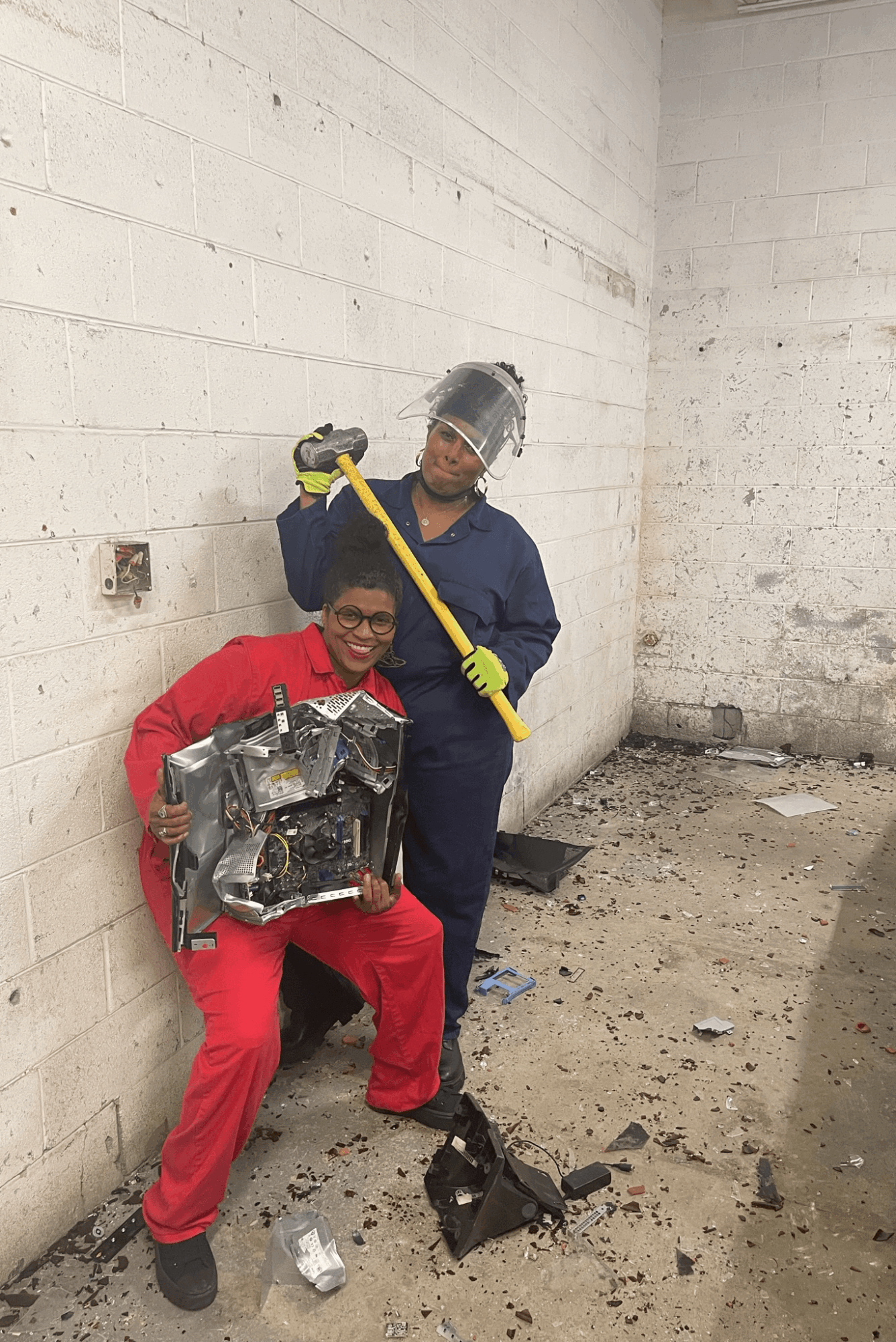 Two workers in protective gear inside a concrete room, one holding a metal object, the other with a sledgehammer.