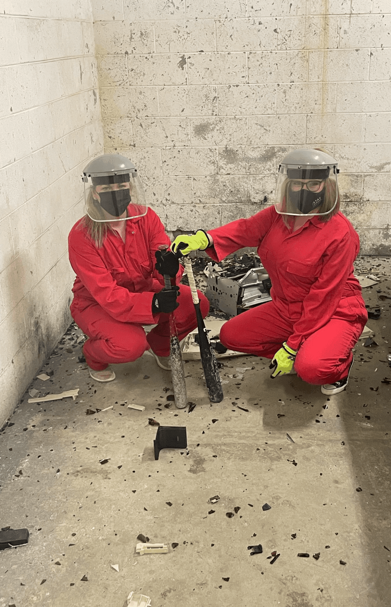 Two workers in red protective suits, face shields, and yellow gloves kneel on a concrete floor in a corner, handling equipment.