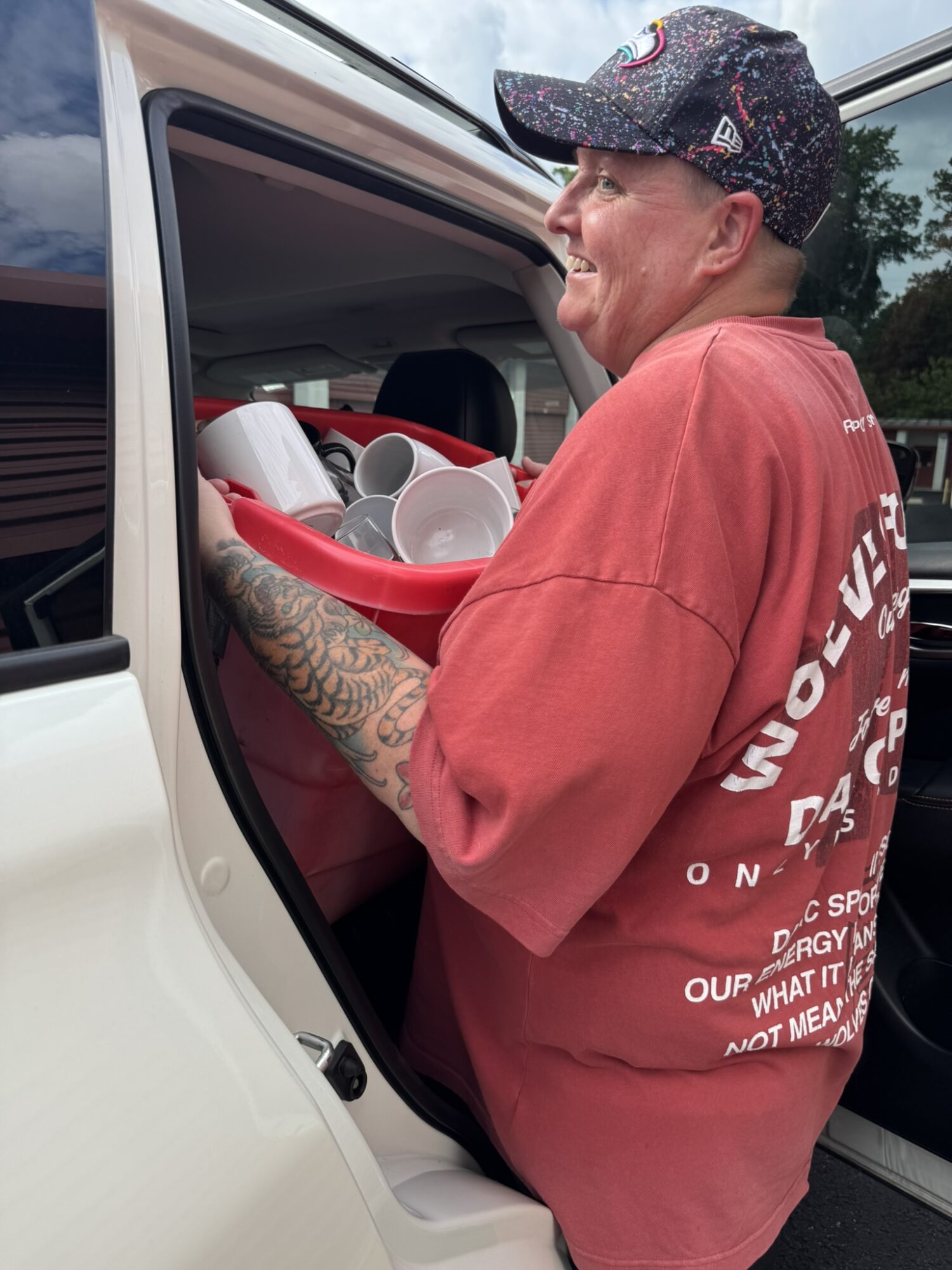 Person with tattoos on arm wearing a red shirt and cap, smiling, holding a tray with cups, standing outside vehicle.