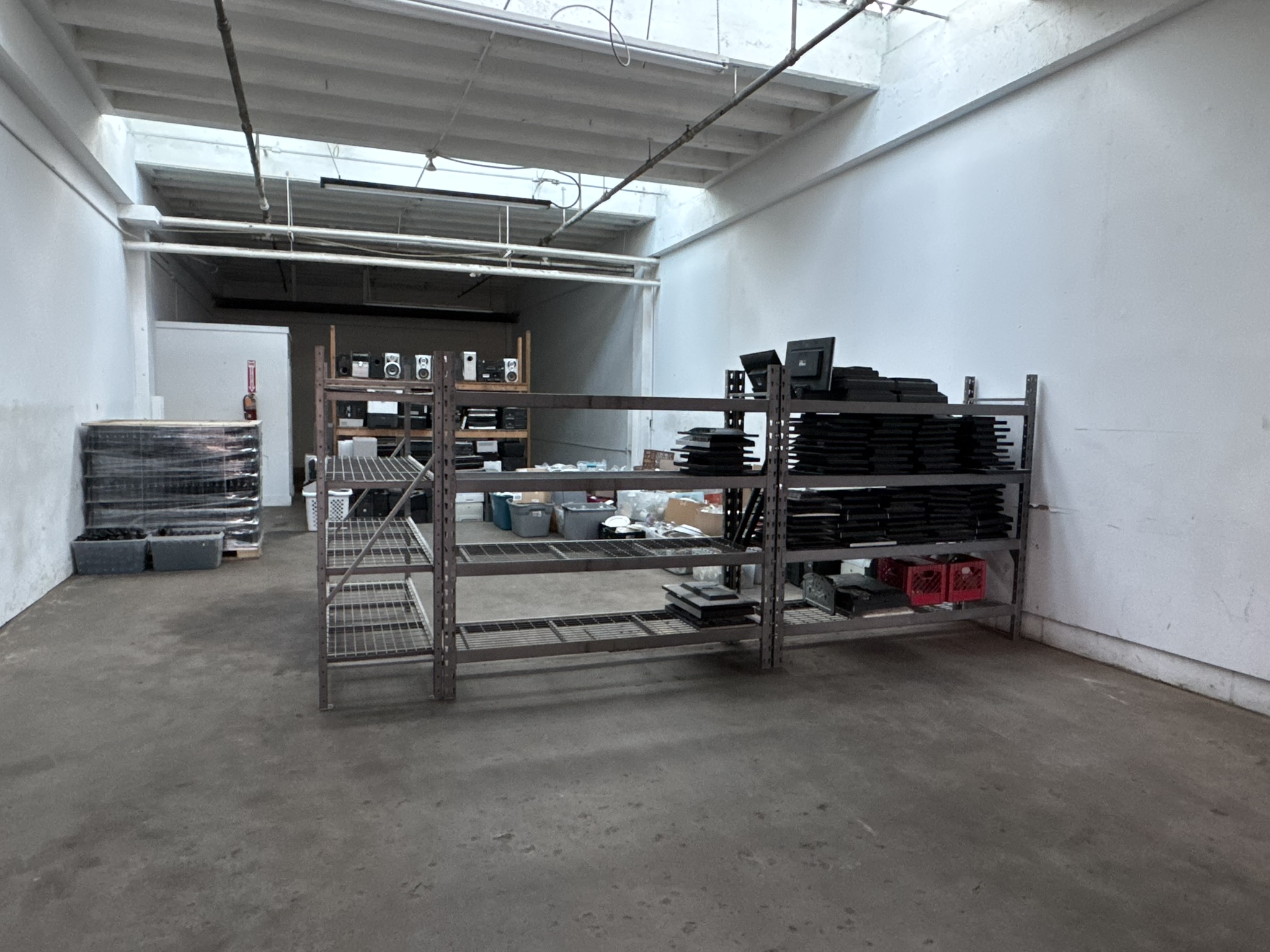 Empty warehouse with metal shelving units and stacked black trays, concrete floor, white walls, and skylight ceiling.