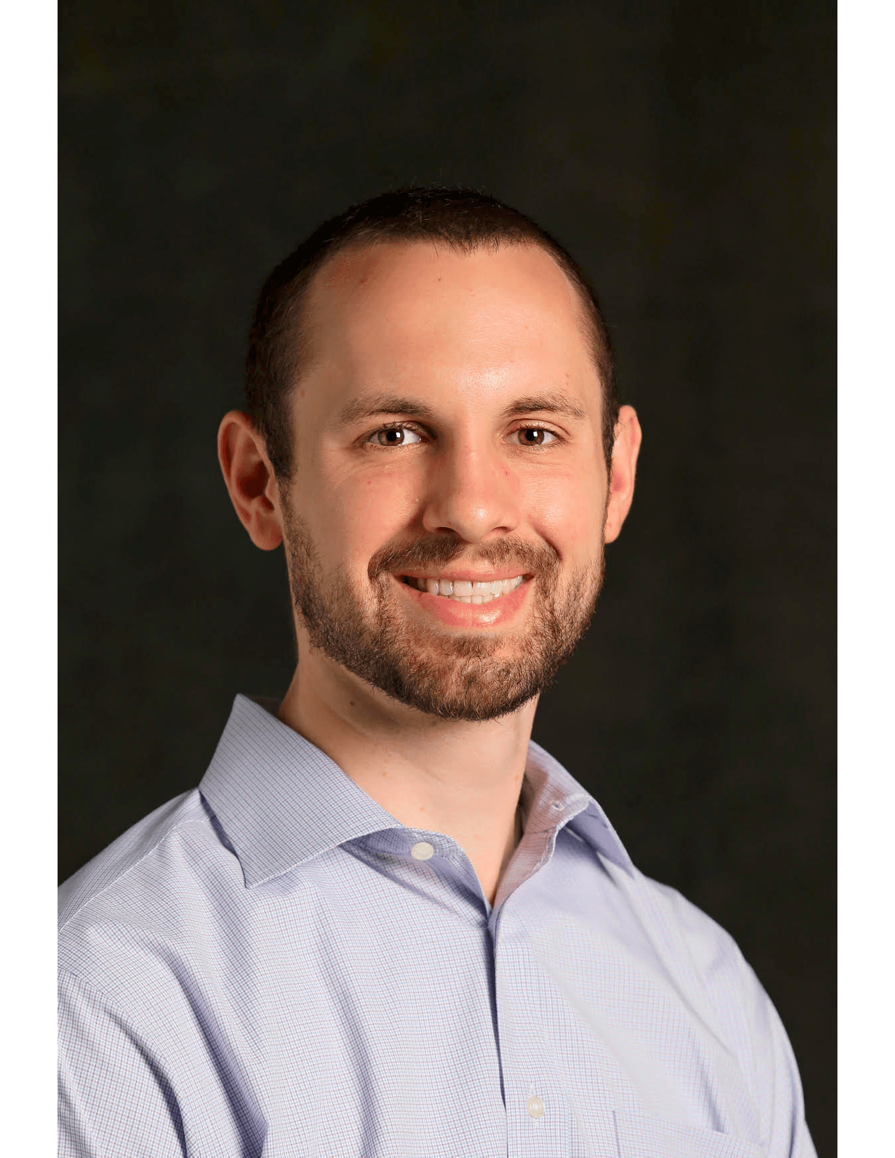 Portrait of a smiling man with short hair and a beard, wearing a light-colored collared shirt, against a dark background.