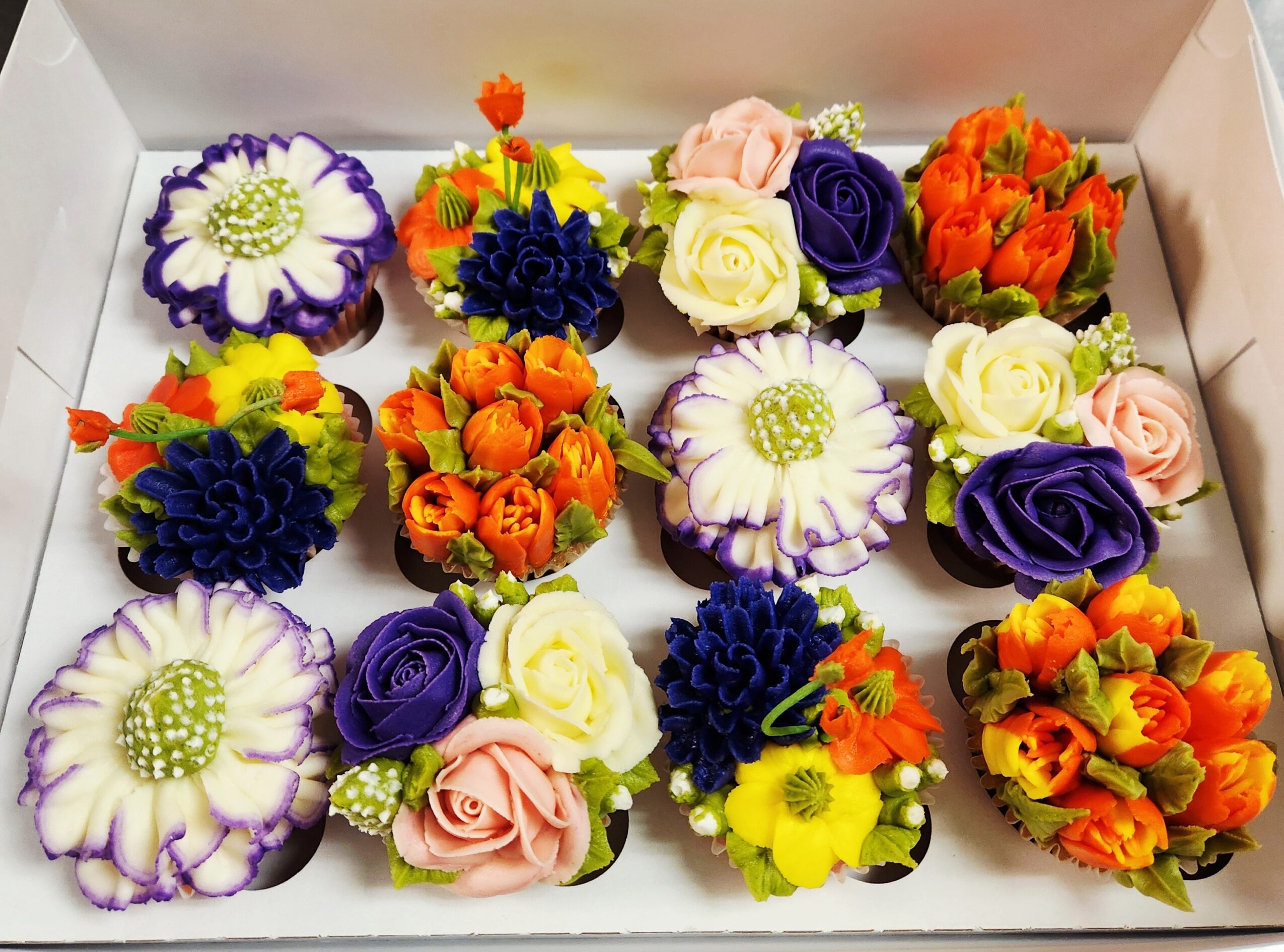 A box of colorful cupcakes decorated with various flower-like frosting designs, including purple, yellow, white, orange, and blue flowers.