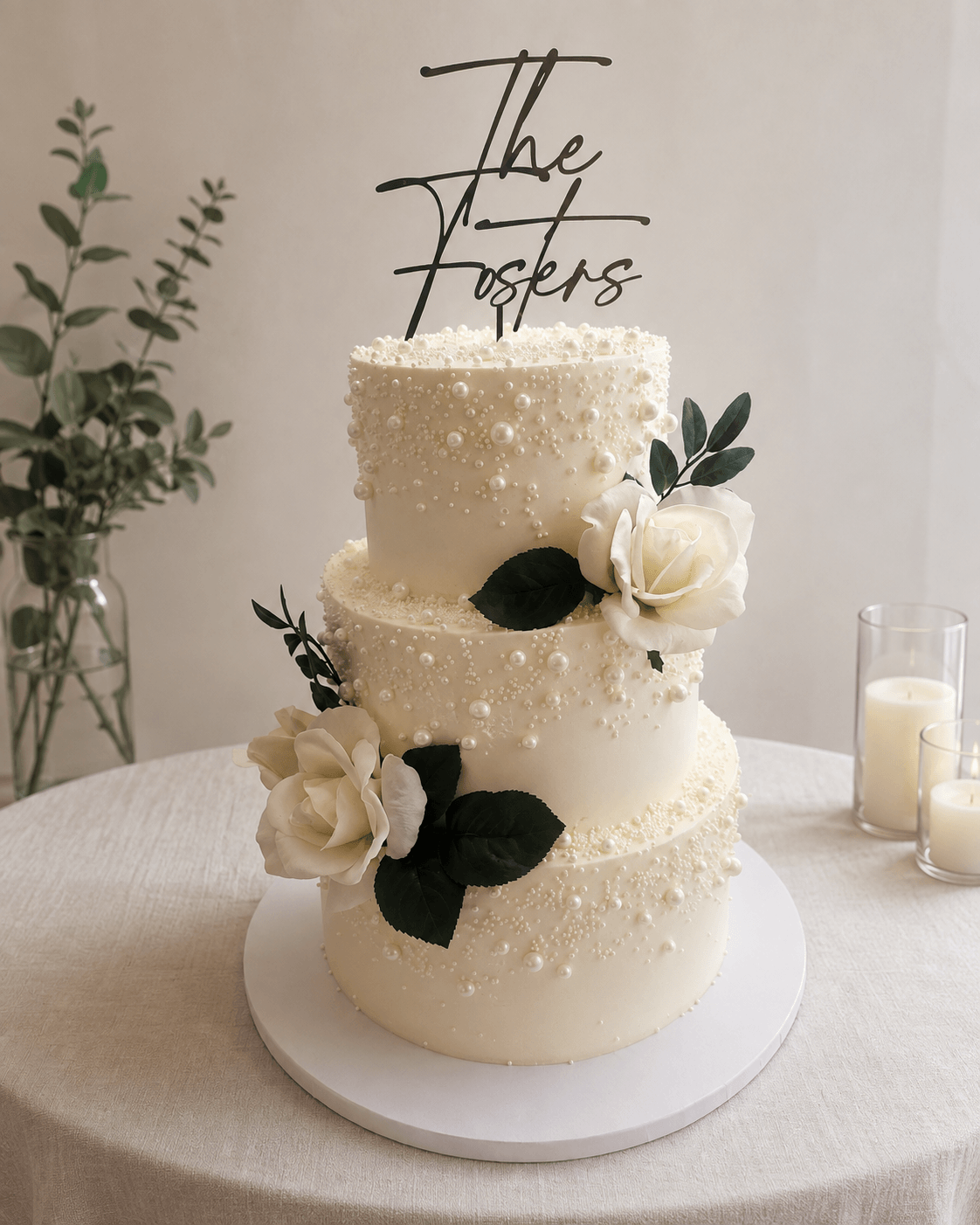 Three-tiered white wedding cake decorated with white roses and dark green leaves, topped with a black script cake topper reading 'The Foster's', on a table with candles and greenery in the background.
