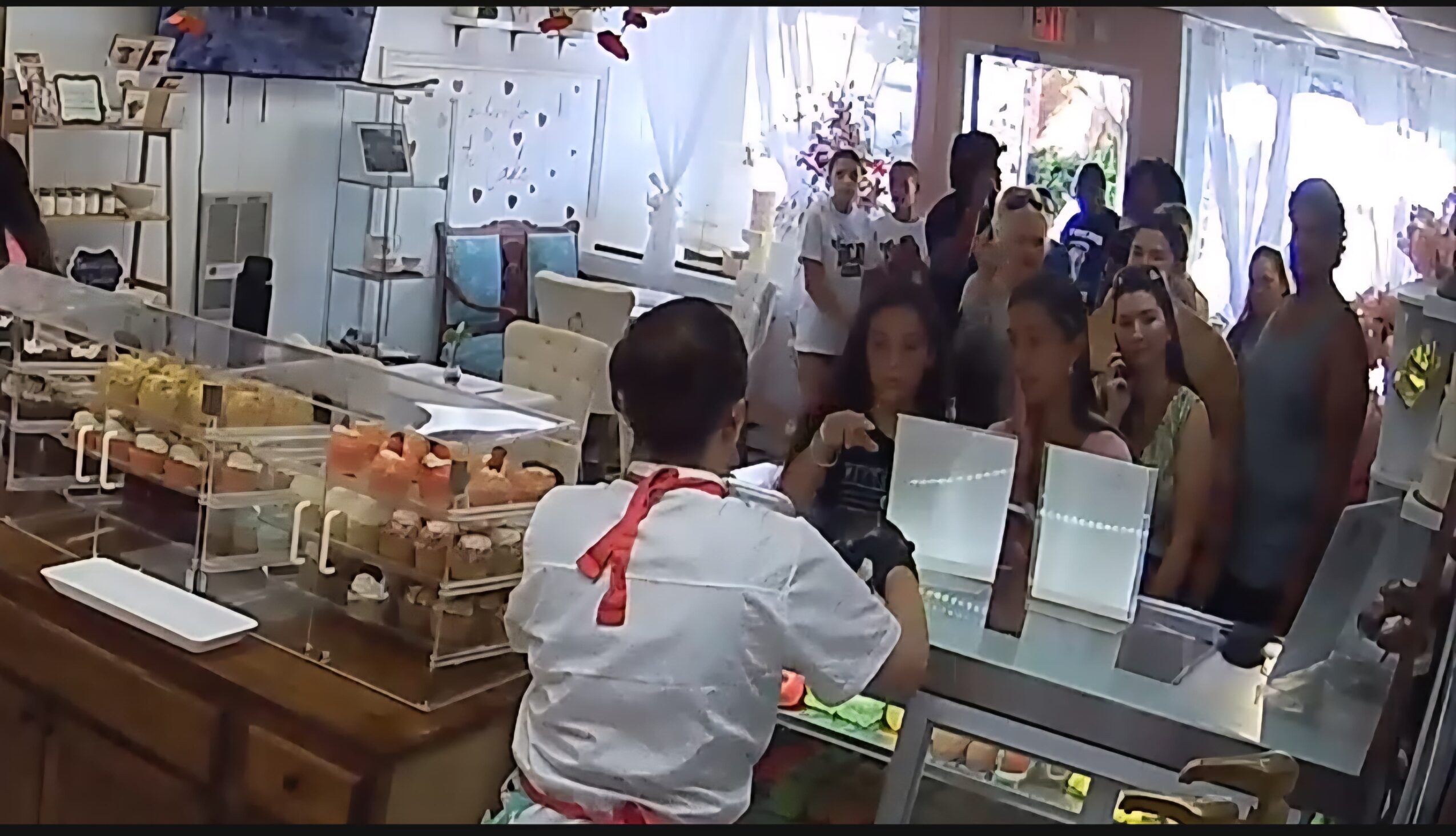 Person in white uniform with red apron serving customers at a counter in a busy bakery or cafe, with people waiting in line.
