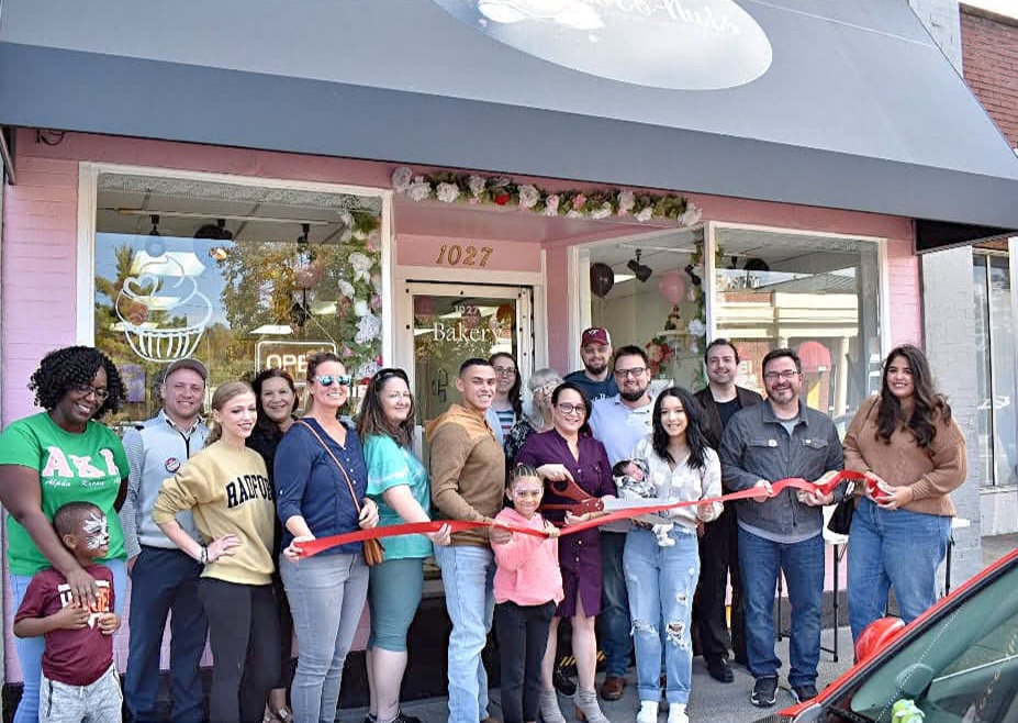 Group of people gathered outside a storefront for a ribbon-cutting event, holding a red ribbon, in front of a pink building.