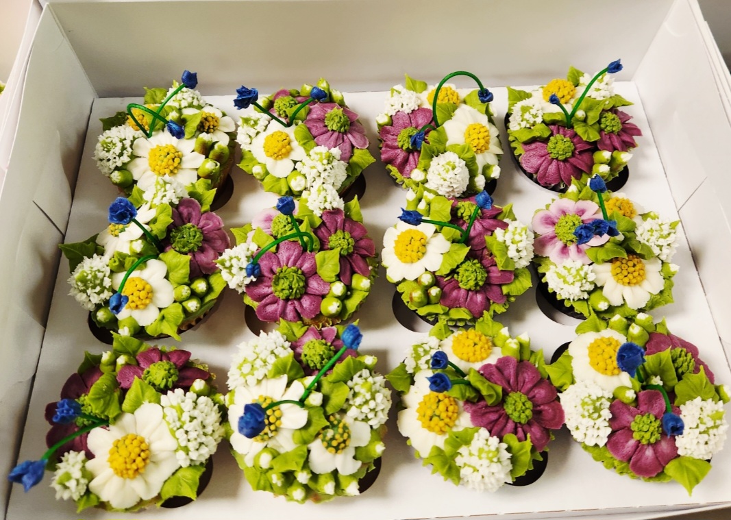 Box of decorated cupcakes with colorful flowers and berries, arranged in three rows, with white, purple, yellow, and green decorations.
