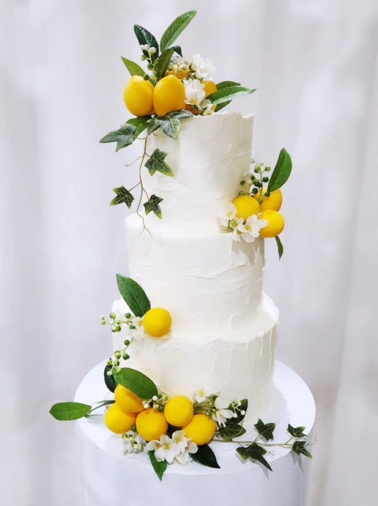 Three-tier white cake decorated with yellow lemons, green leaves, and white flowers, set against a white background.