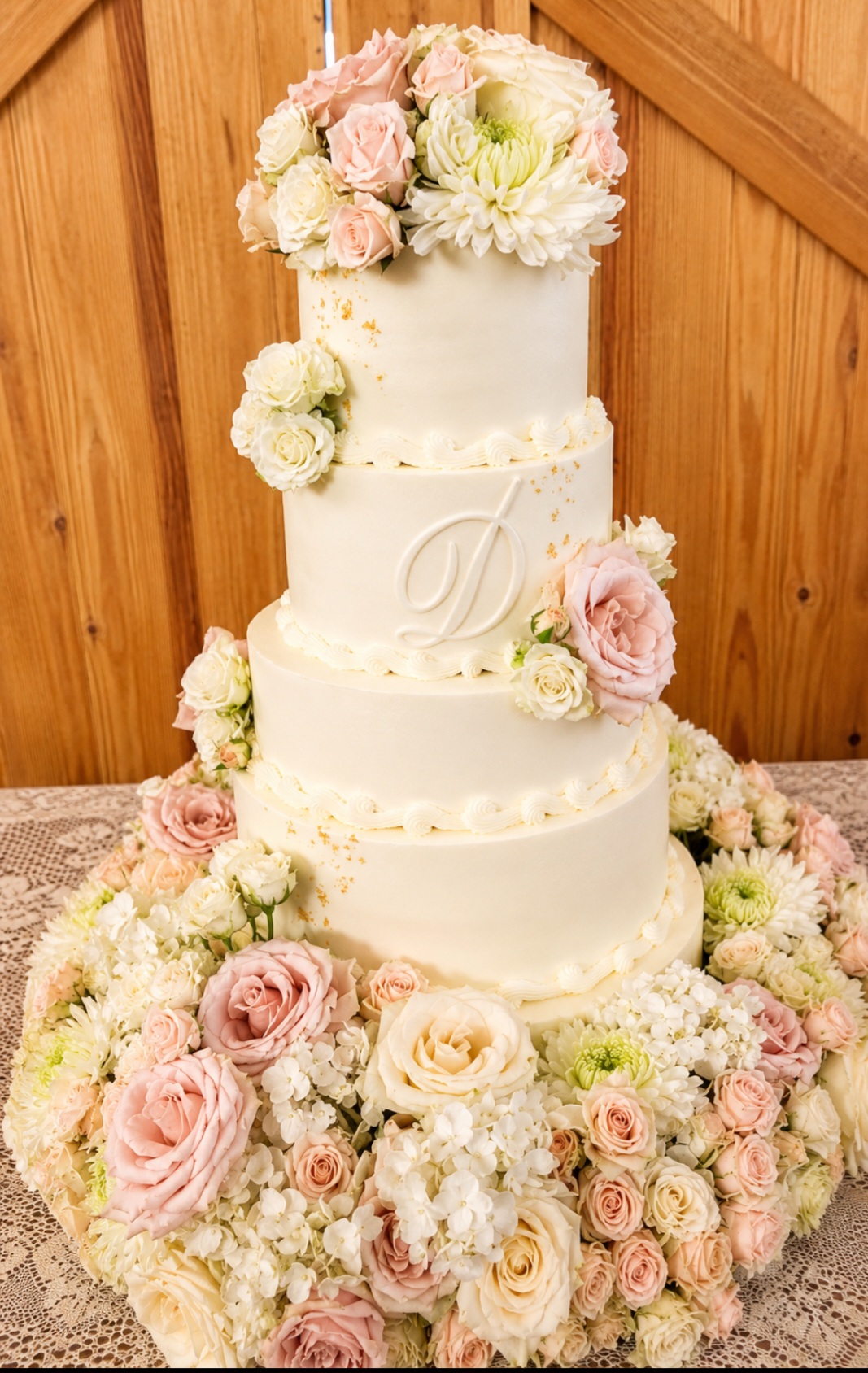 Four-tier white wedding cake decorated with pink and white flowers, surrounded by a floral arrangement on a table.