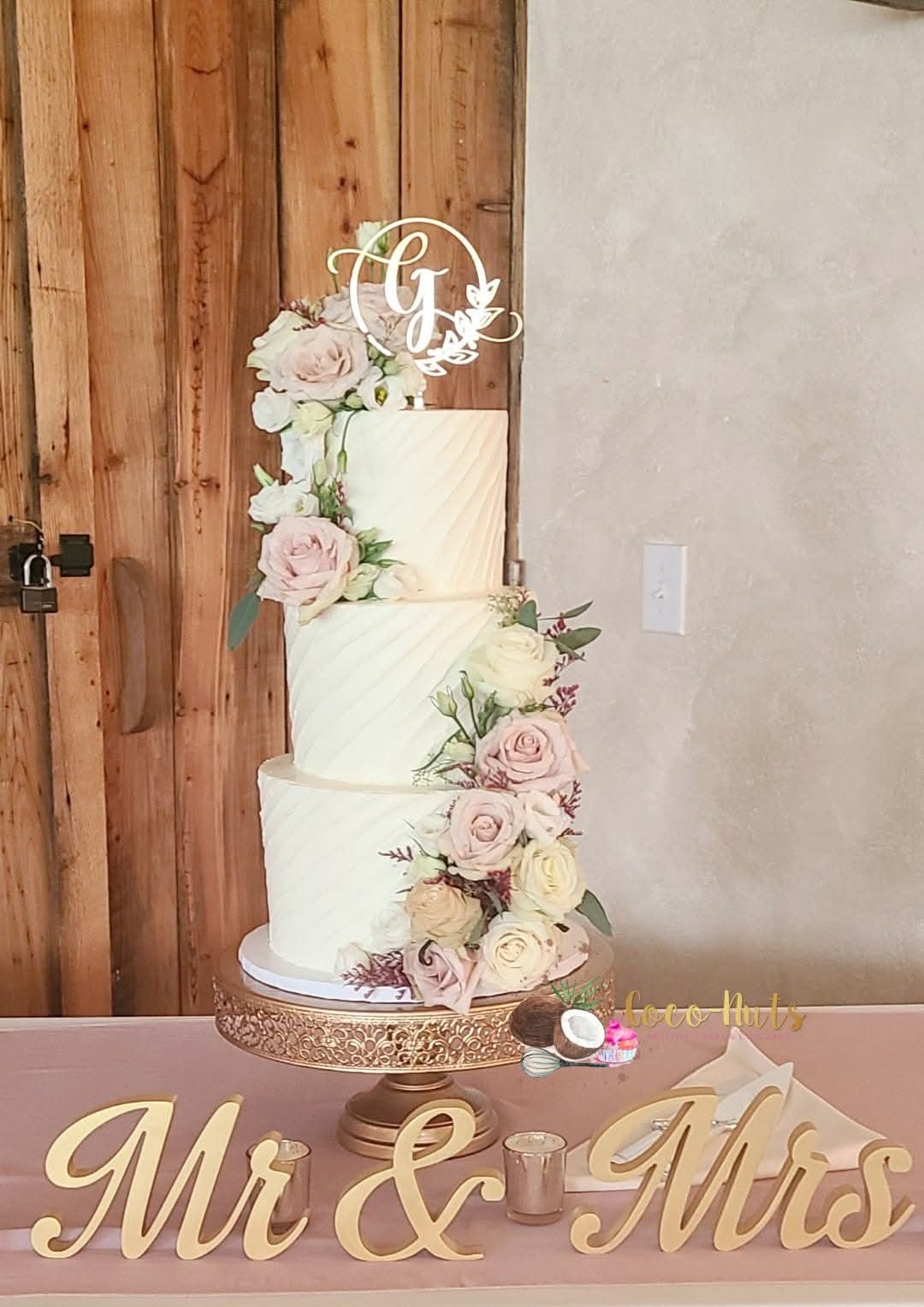 Four-tier wedding cake decorated with pink and white flowers, topped with a monogram cake topper, on a gold stand.