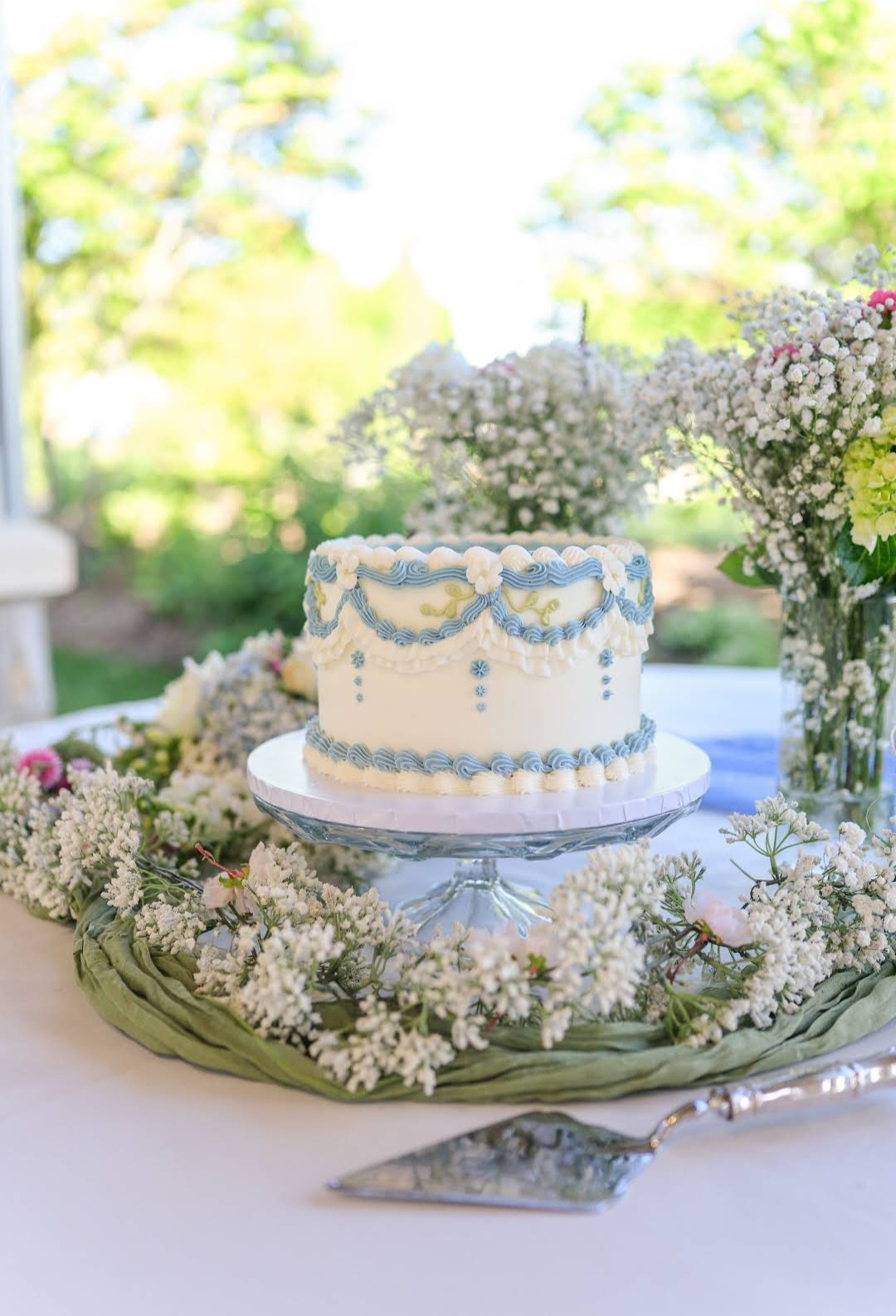 Decorative cake on a table surrounded by white flowers and greenery, outdoors with sunlight and trees in background.
