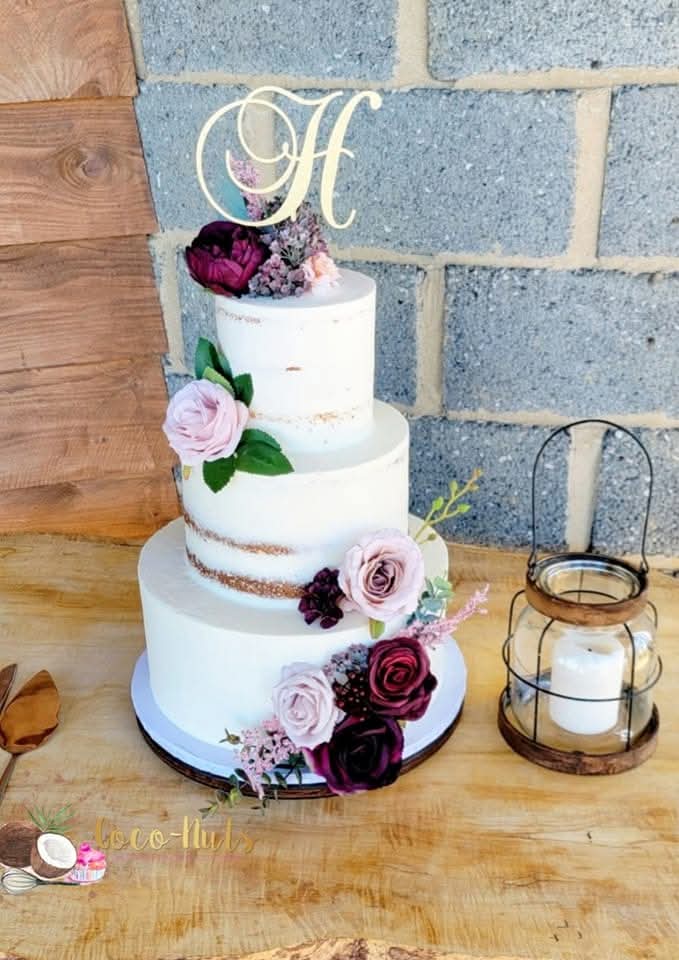 Three-tier white wedding cake decorated with pink and purple flowers, topped with a gold letter 'H', on a wooden table.