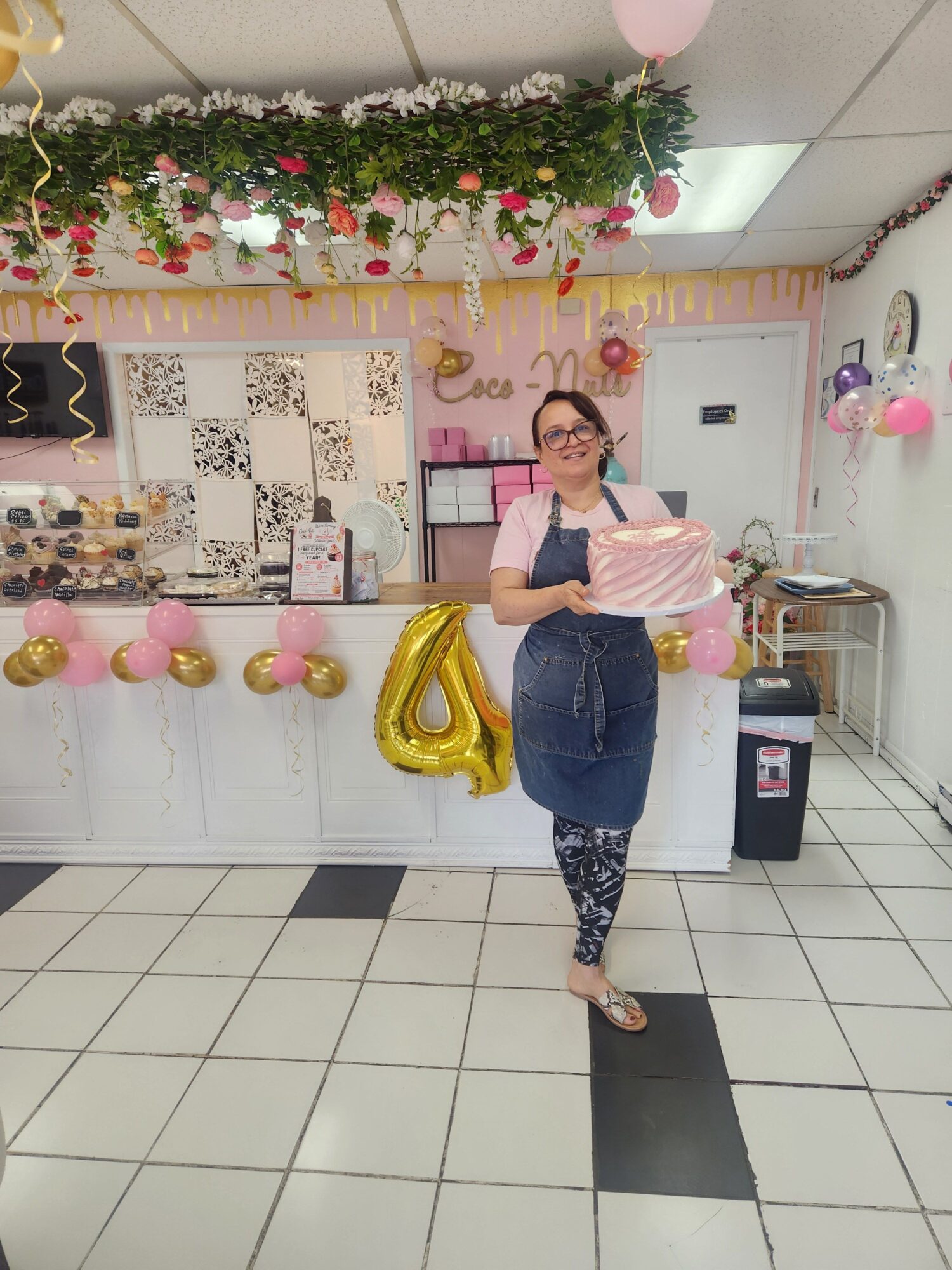 Person standing in a decorated room with balloons, flowers, and a large gold number 1 balloon, taking a selfie.