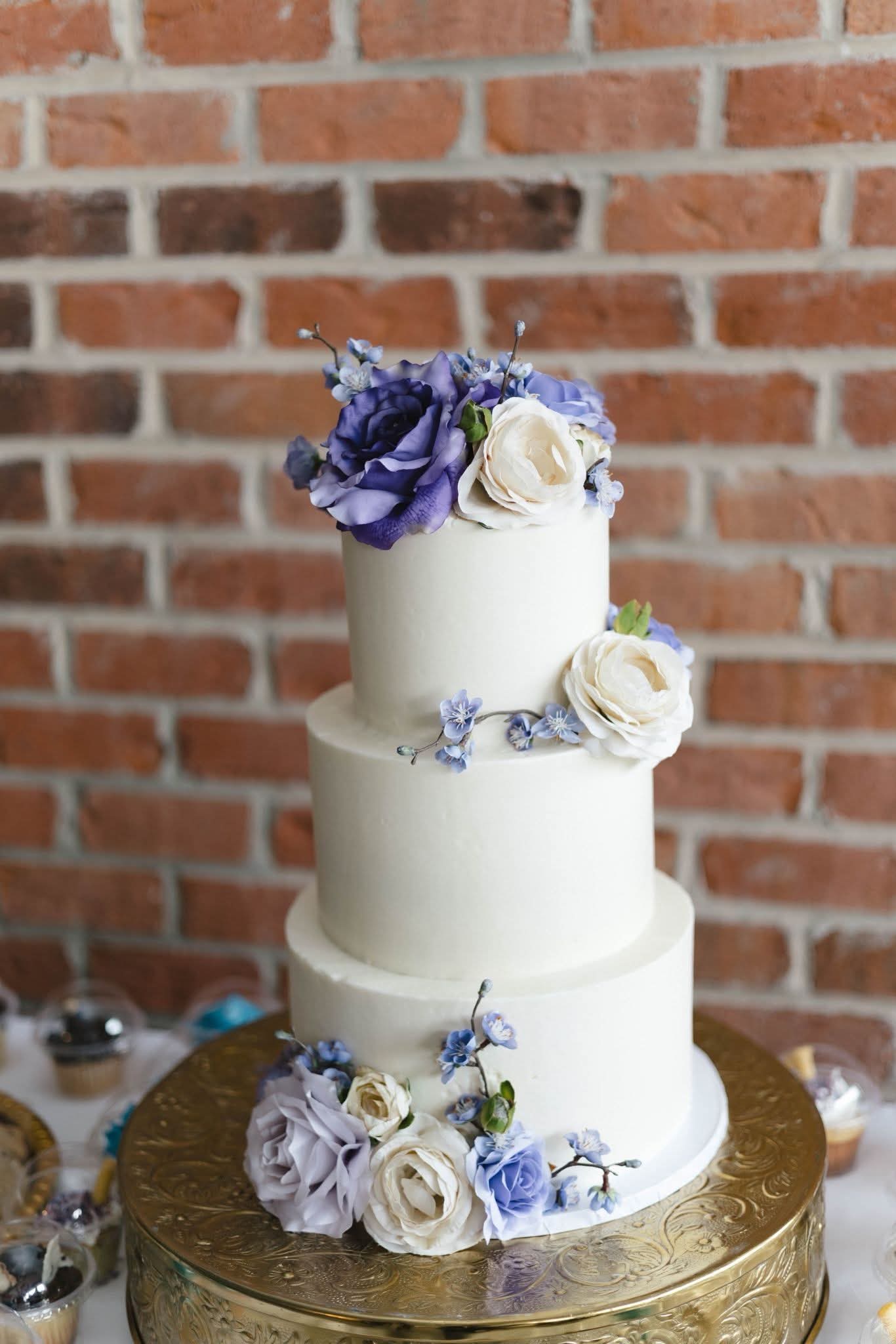 Three-tiered white wedding cake decorated with purple and white flowers, on a gold cake stand, against a brick wall.
