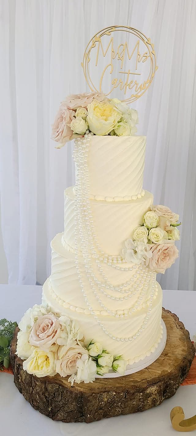 Four-tiered white wedding cake decorated with flowers on a wooden base, with a gold topper reading 'Mr. & Mrs.'