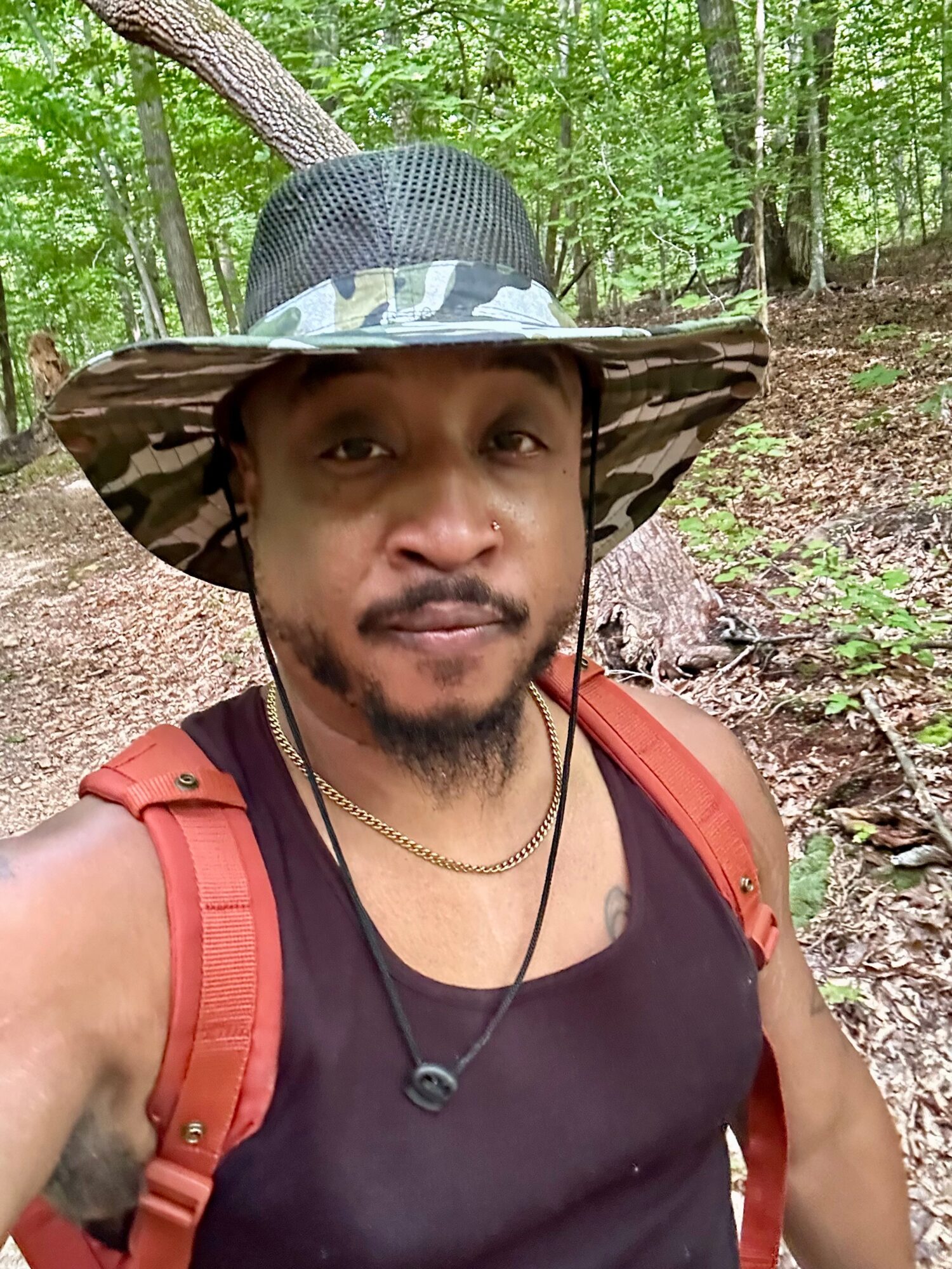 Man wearing a wide-brimmed hat, black tank top, and red backpack in a forest, looking at the camera.