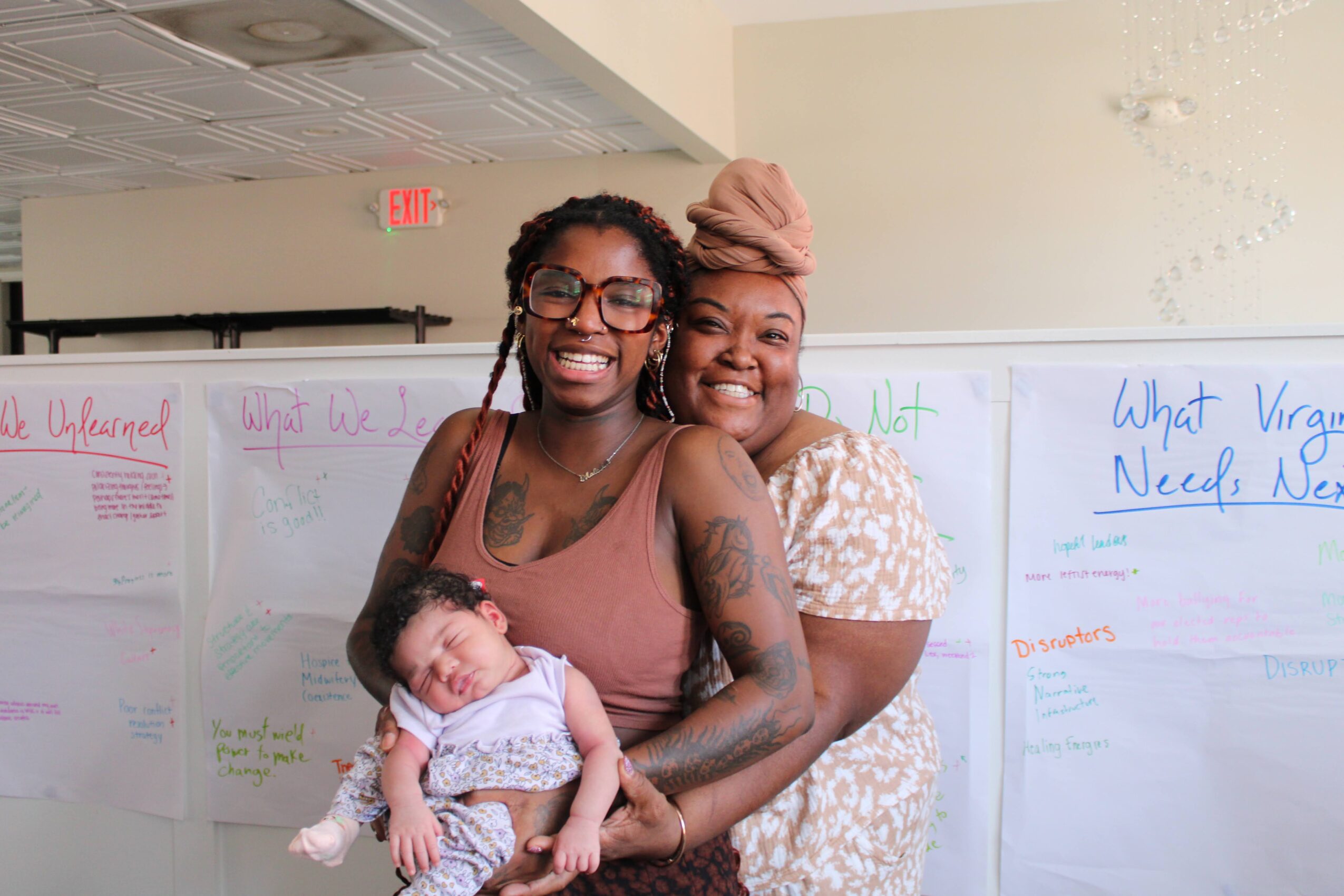 Two women smiling and holding a baby in front of whiteboards with handwritten notes, in a classroom setting.