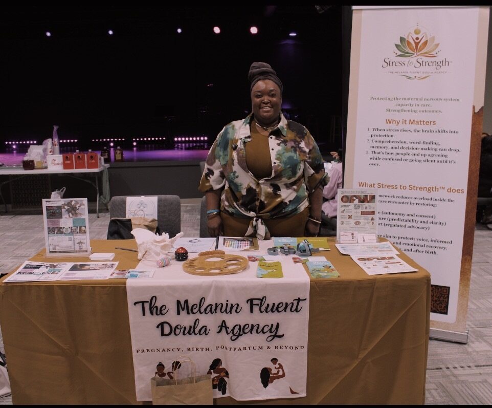 Woman standing behind a table with promotional materials at an event, with a banner and a sign for The Melanin Fluent Deula Agency.