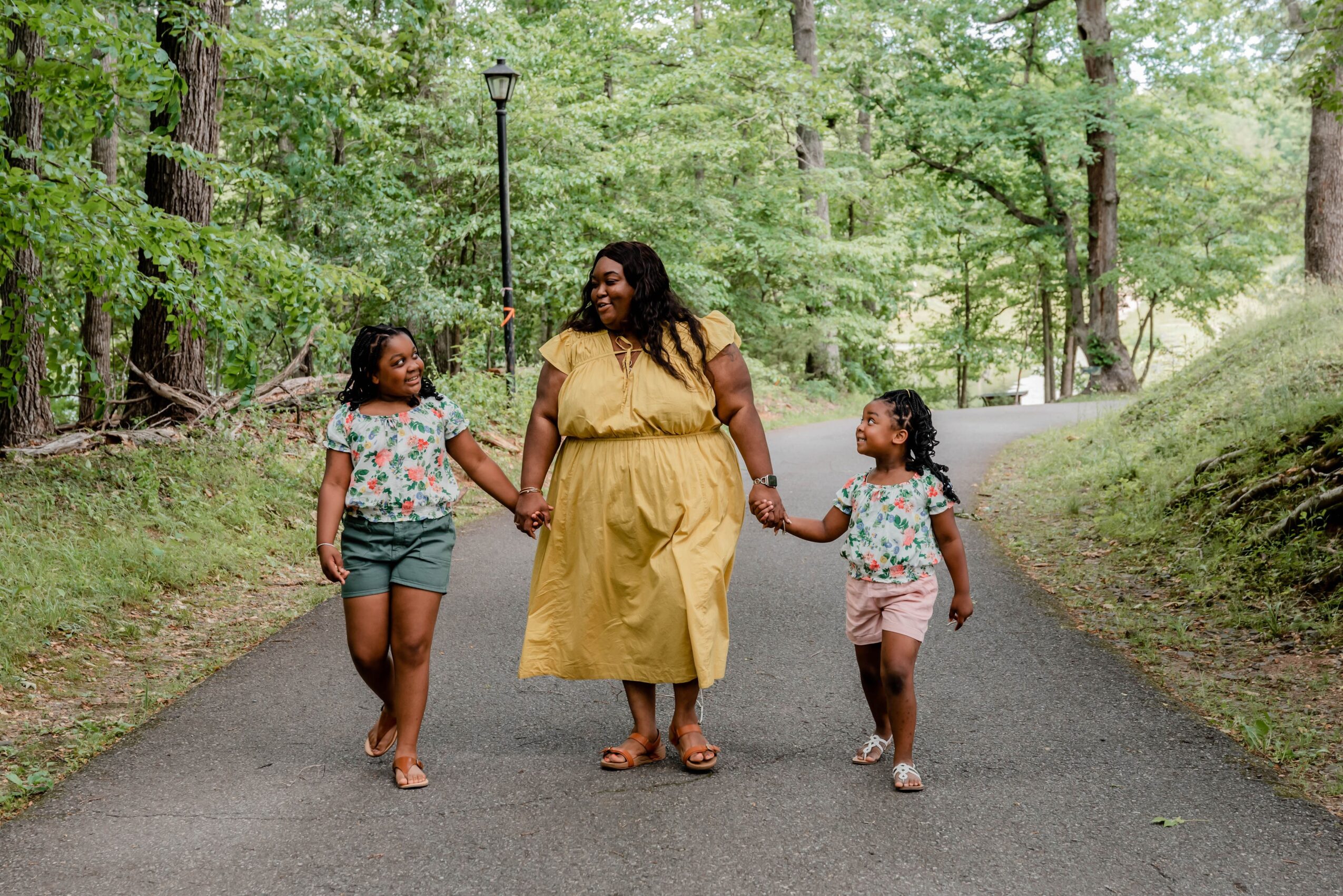 Woman walking with two children on a paved path in a wooded park, holding hands, surrounded by green trees.