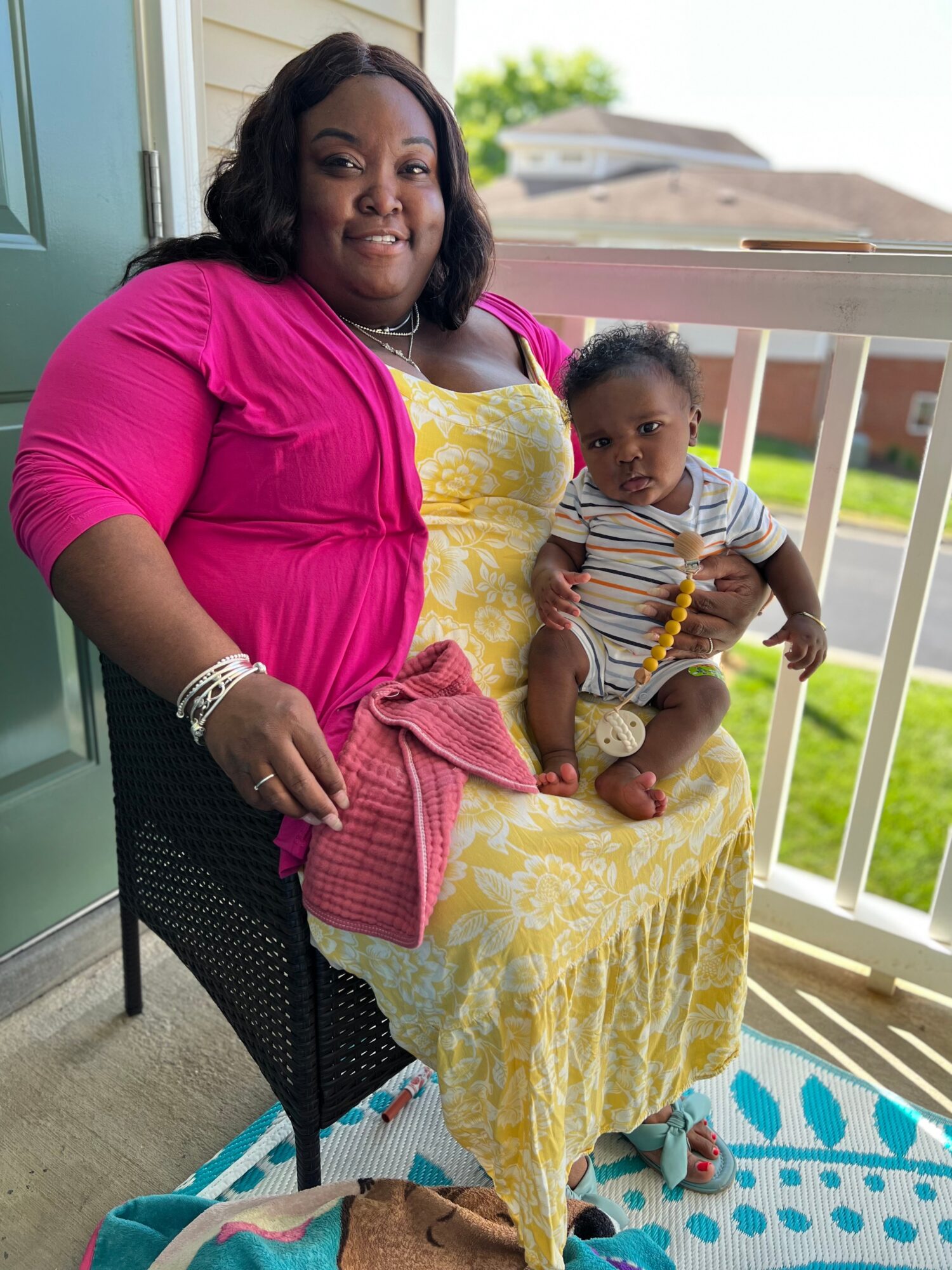 Woman sitting on a chair holding a young child on her lap on a porch with a railing and yard in background.