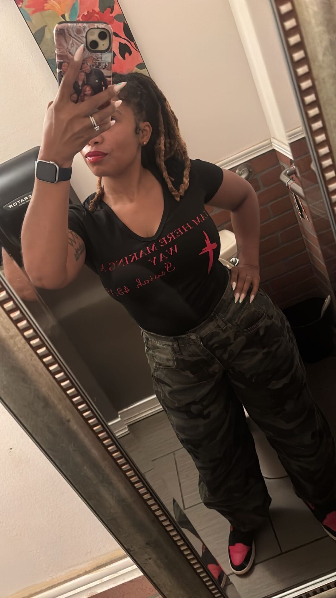 Woman taking a mirror selfie in a bathroom, wearing a black shirt and camouflage pants.