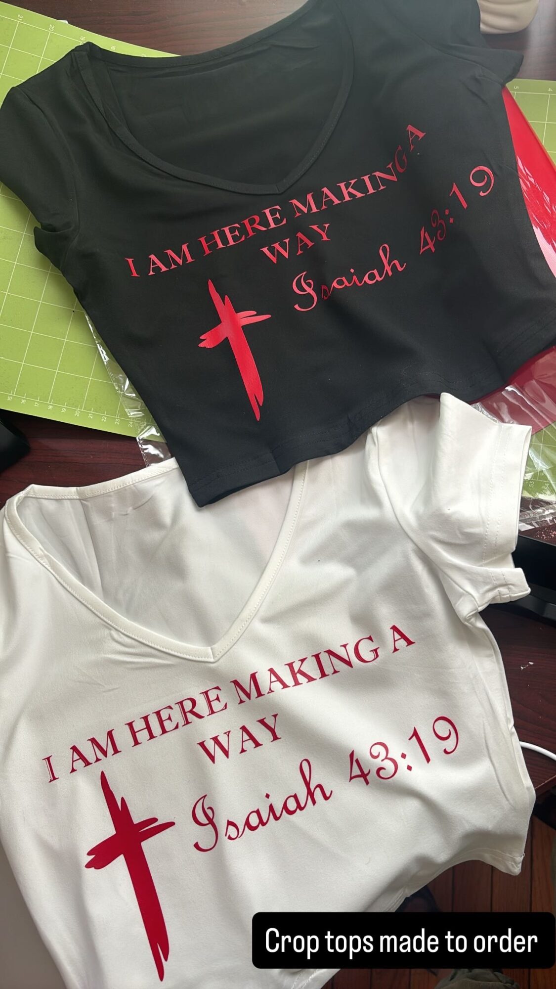 Two T-shirts, one black and one white, with red text and a cross, laid on a surface. Black shirt says, 'I am here making a way,' white shirt says, 'I am here making a way,' both referencing Isaiah 43:19.