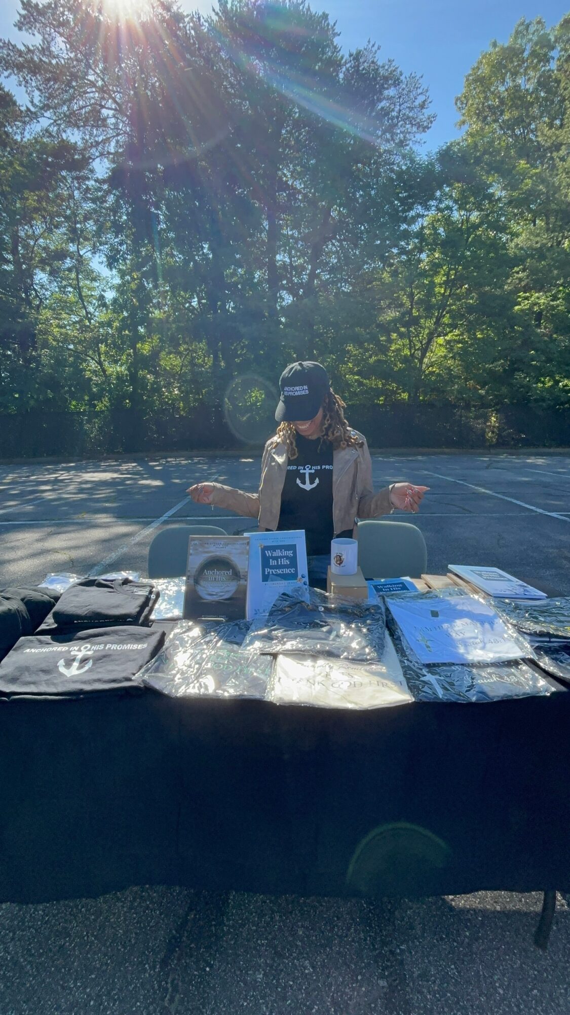 Person wearing a cap standing behind a table with various items outdoors, trees and sunlight in background.