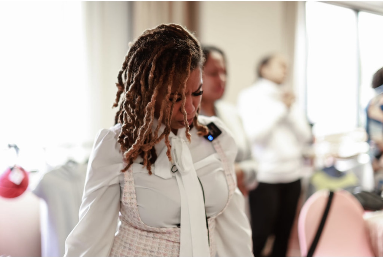 Woman with dreadlocks looking down in a bright room with people in the background.