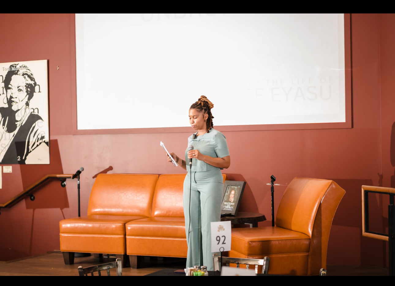 Woman in light blue dress standing on stage, holding a paper, with orange sofa and framed picture behind her.