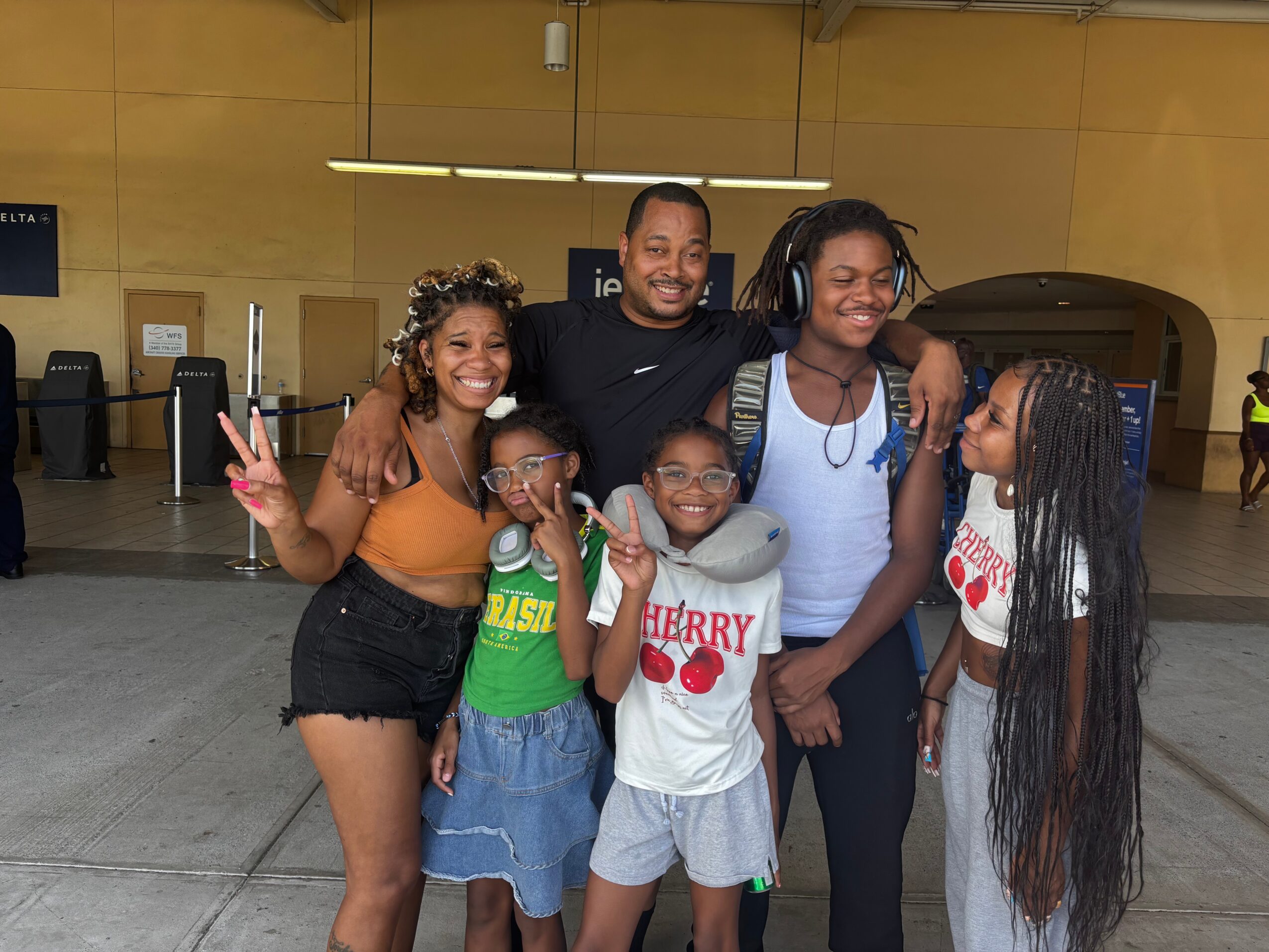 Group of five smiling people posing indoors, some making peace signs, with a yellow wall background.