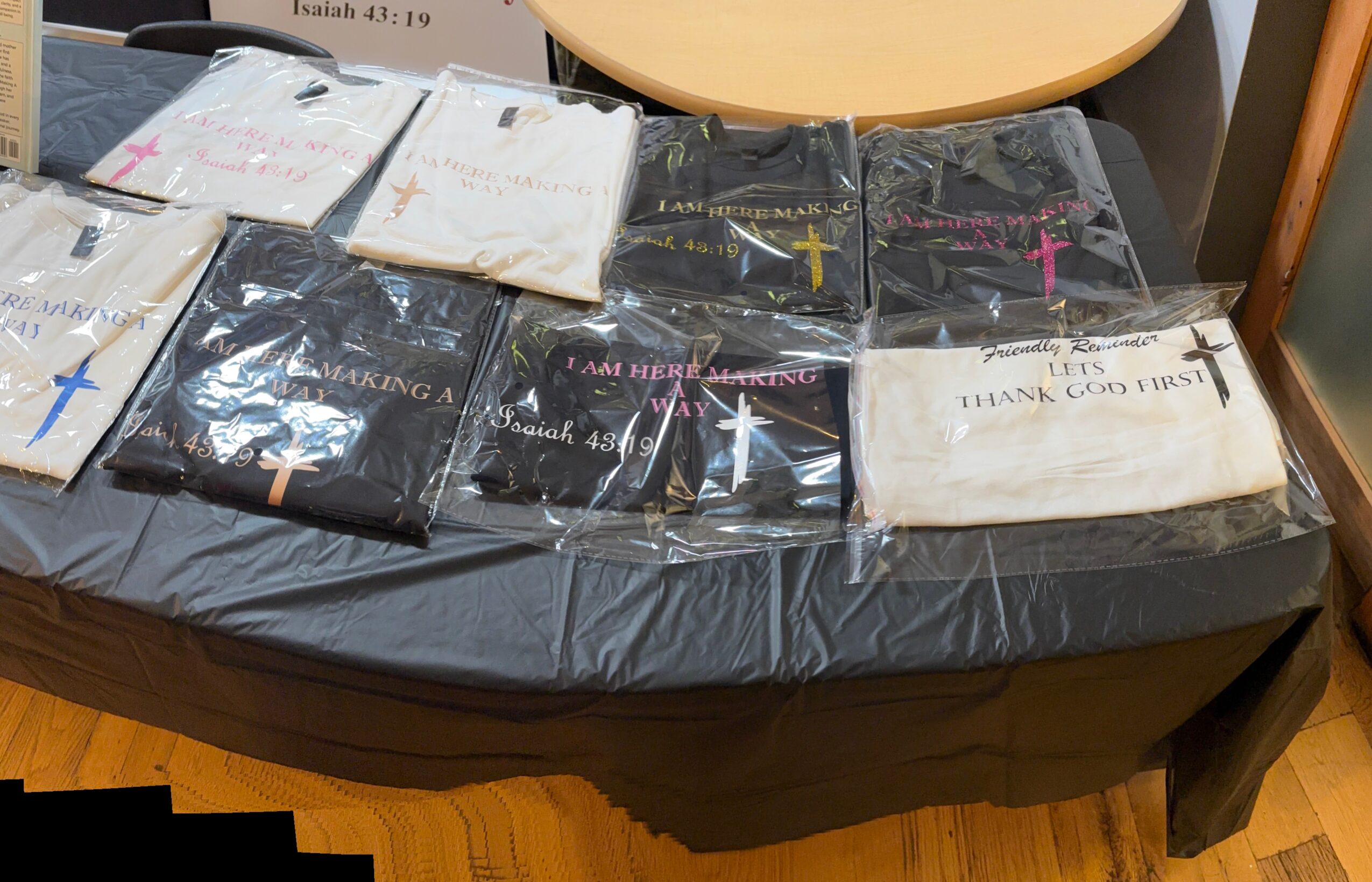 Table covered with folded cloths, some with crosses, in plastic packaging, and a paper bag, in a room with wooden flooring.